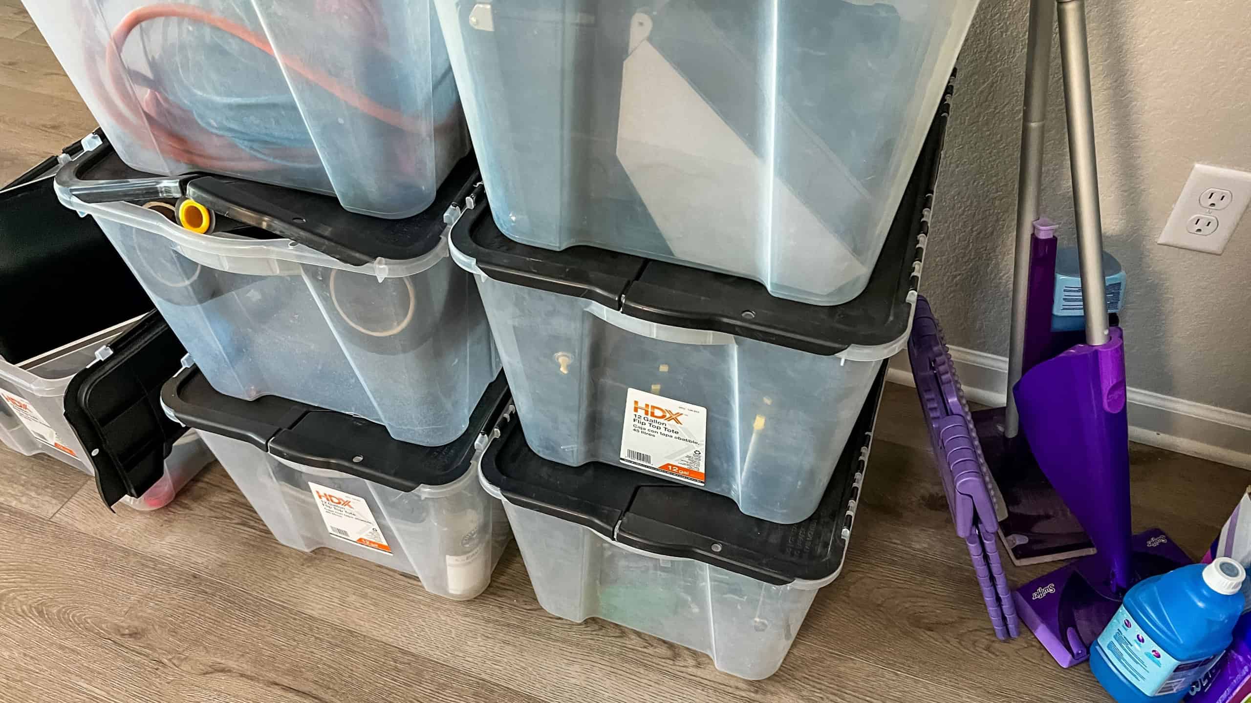 A collection of clear plastic storage bins with black lids stacked against a wall used for organizing and packing during a moving process