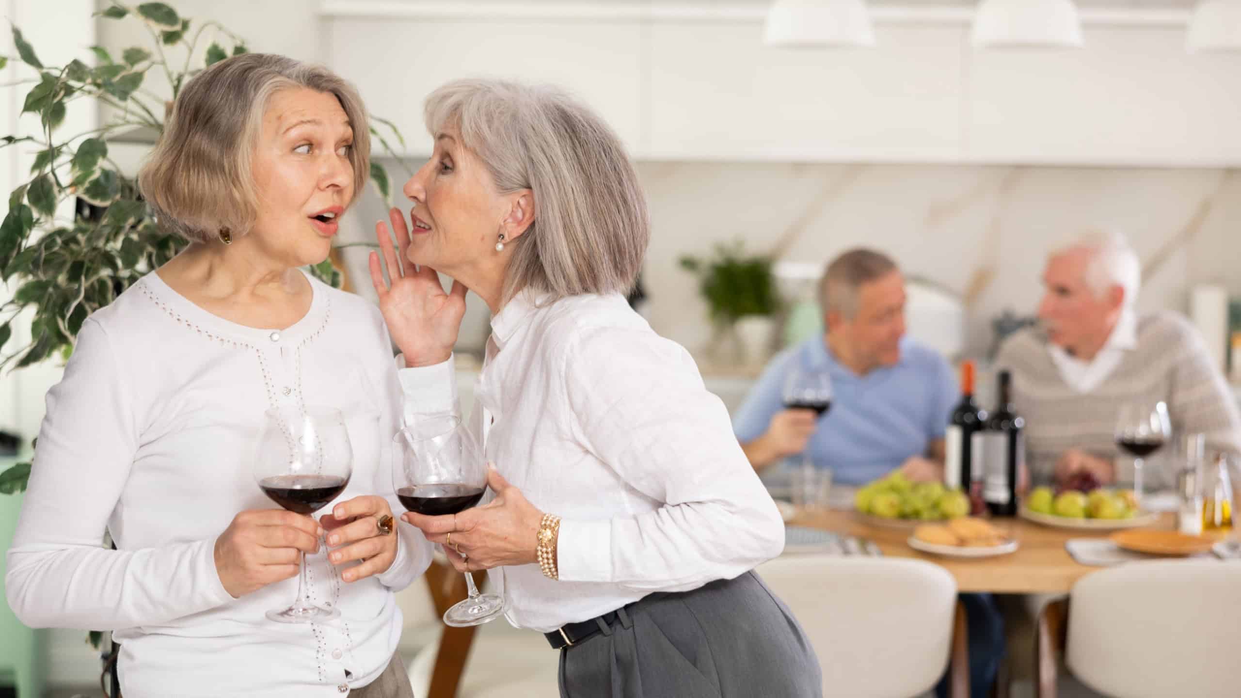 During friendly meeting, two elderly ladies with glass of wine in their hands stand at distance from table, communicate and chatting, smile, keep secrets.