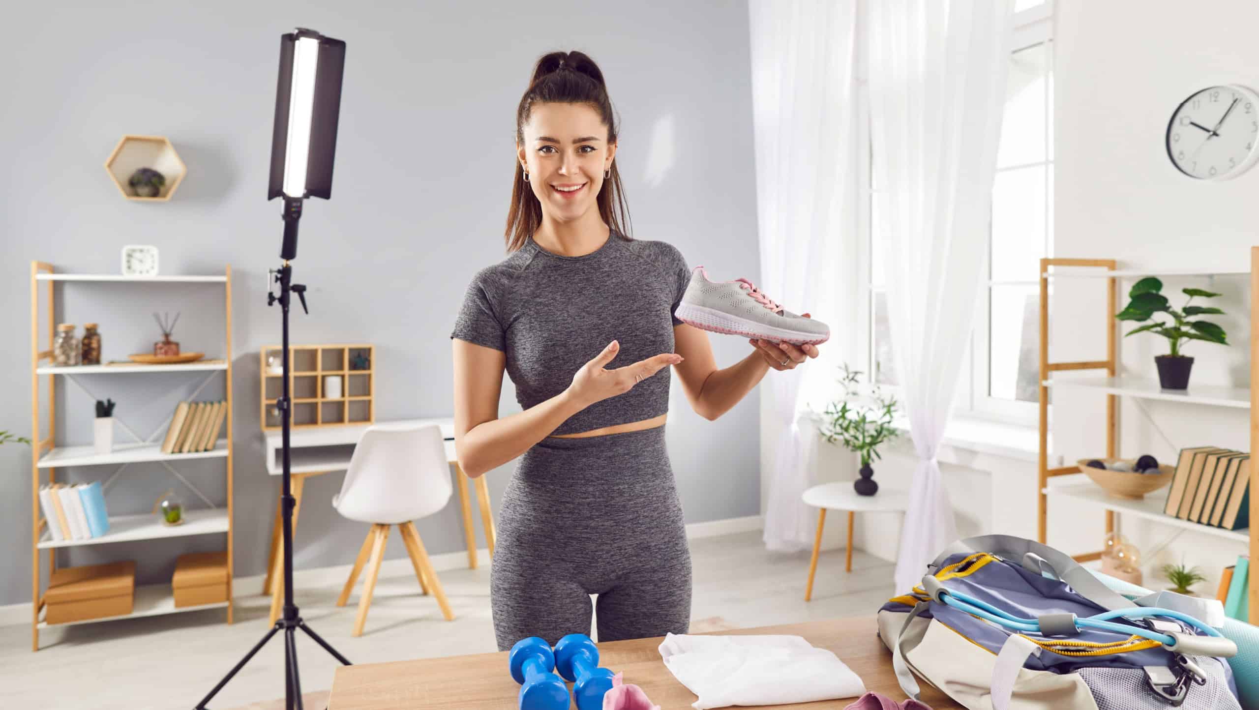 Sporty smiling young woman fitness instructor recording video and showing sneakers preparing stuff for training packing gym bag. Girl sport blogger making video for fitness blog on smartphone.