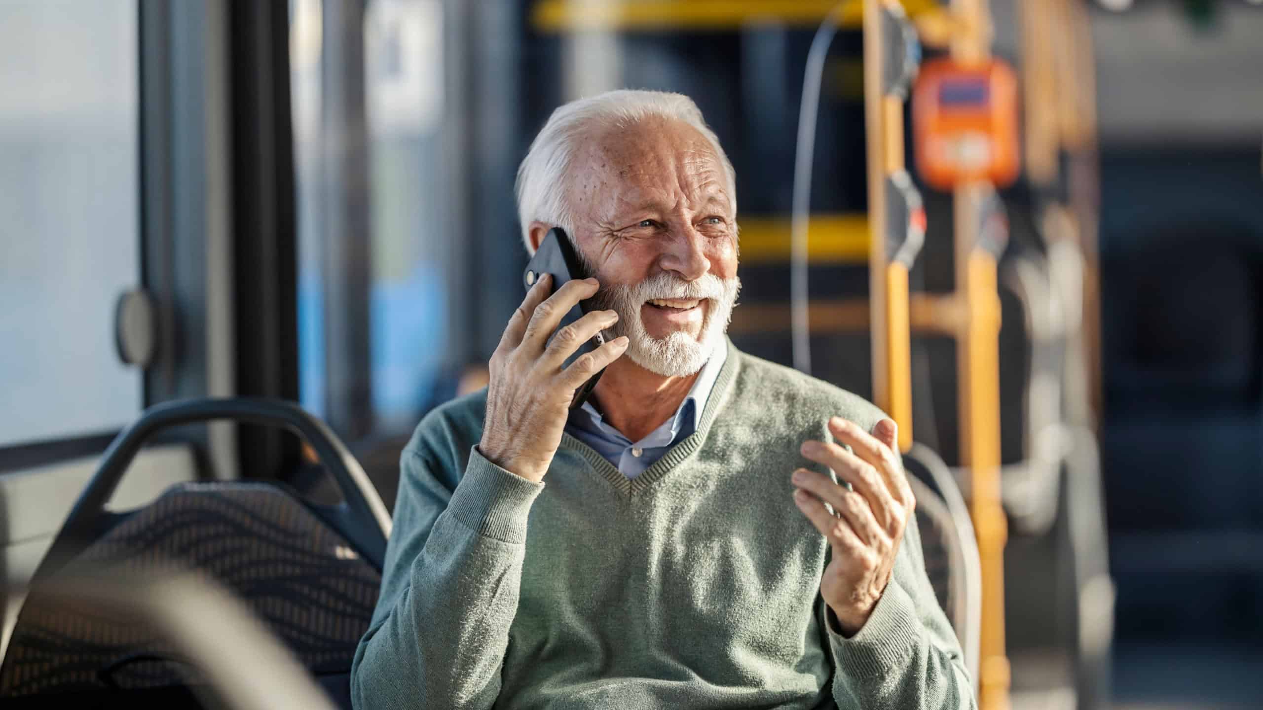 Portrait of happy mature man in smart casual sitting in bus and talking on his phone.