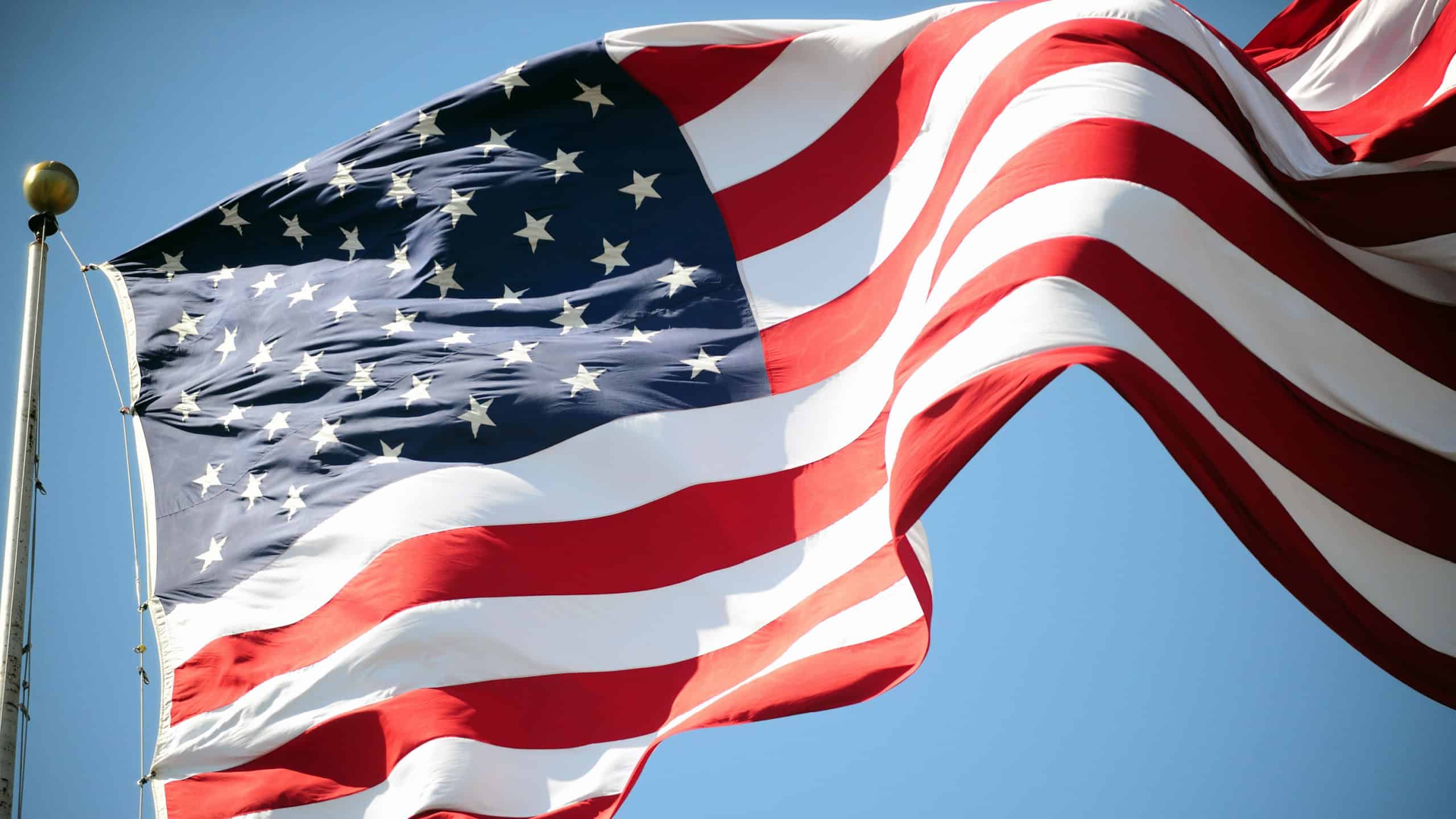 The American flag waves against a clear blue sky. Stars and stripes are prominent on the flag. The flag symbolizes patriotism and American identity.