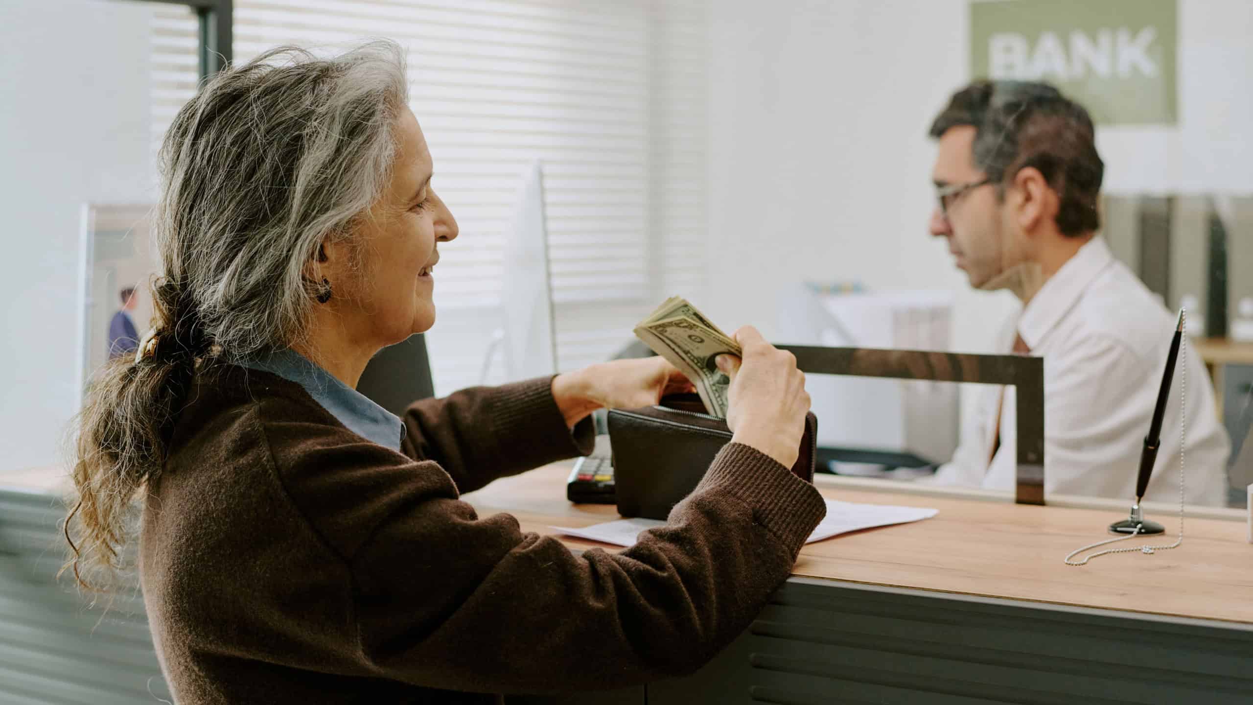 Senior woman handing money to bank teller at service desk during transaction interaction, bank employee assisting customer with financial services