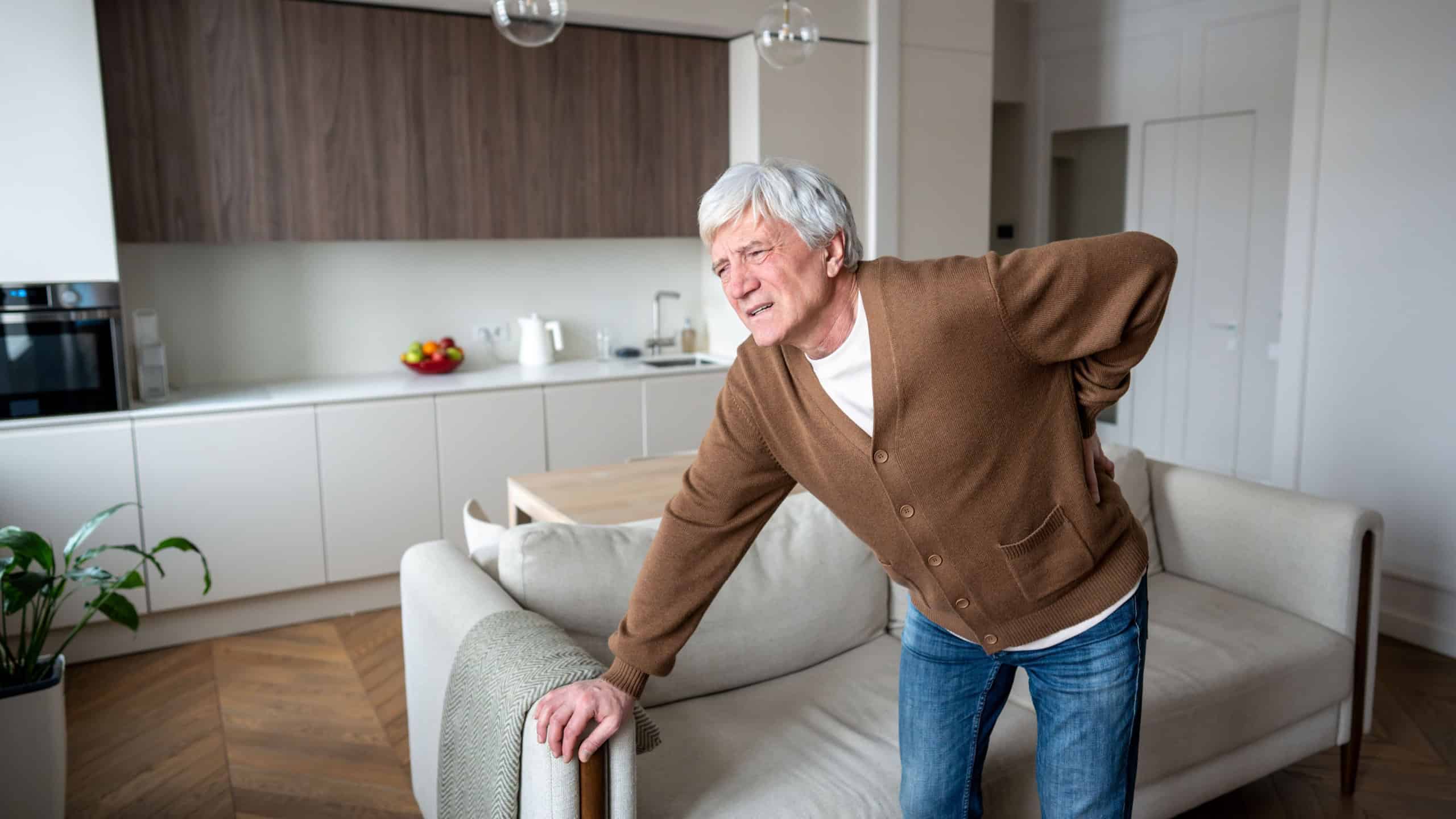 Bent sad unhappy senior man holds painful lumbar back leaning on sofa in silent agony. Degenerative disc deterioration, sciatic nerve inflammation, vertebral collapse, chronic stiffness illness