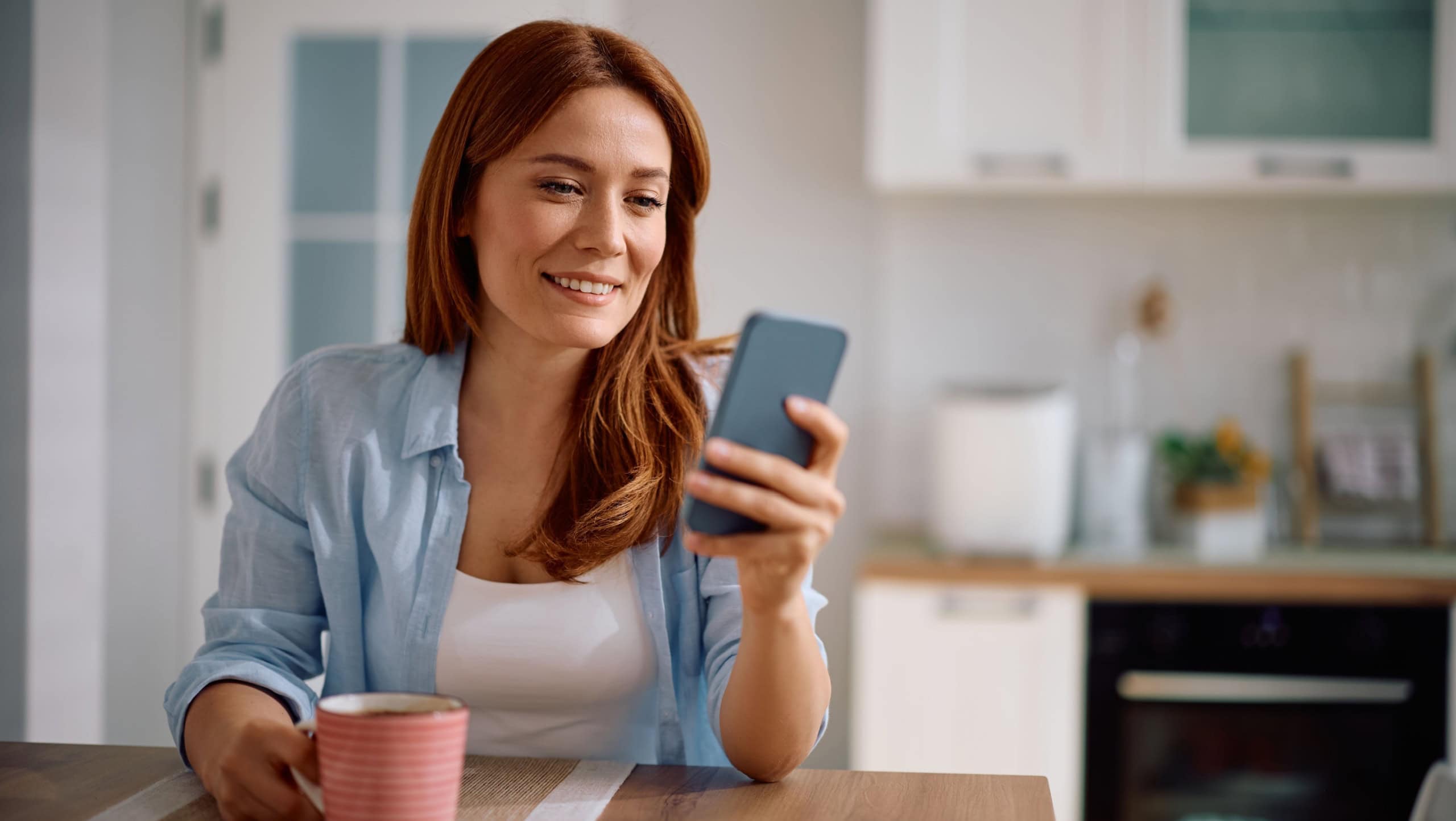 Happy woman drinking coffee while reading text message on mobile phone at home.