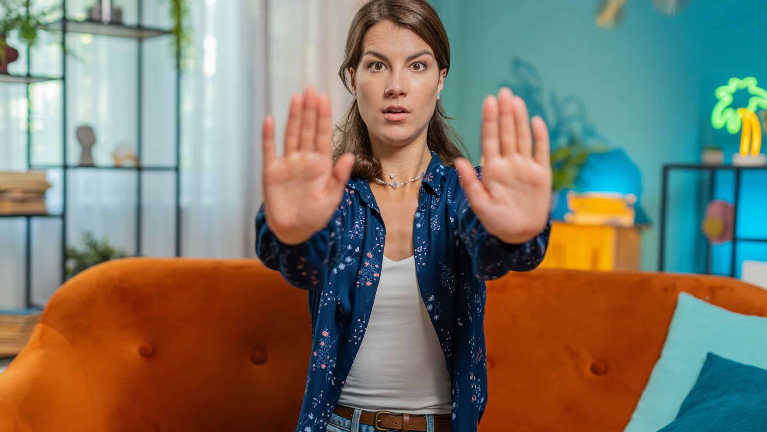 Stop gesture. Angry Caucasian woman says No hold palm hands, warning of finish, prohibited access, declining communication, body language, danger. Girl at home blue room sits on orange sofa couch.