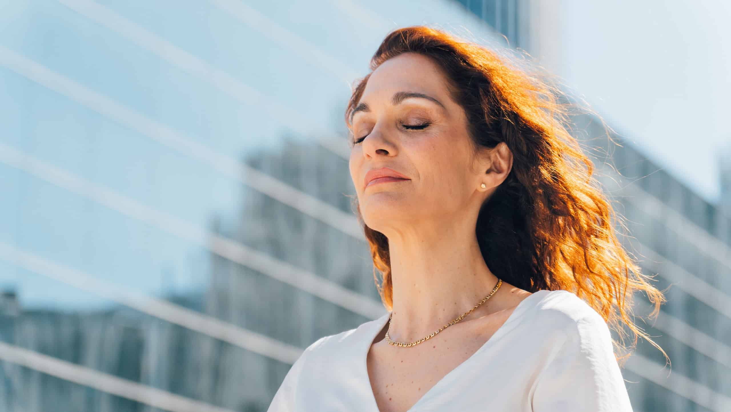 Businesswoman is breathing fresh air in front of modern office building with closed eyes, enjoying her break and improving her mental health