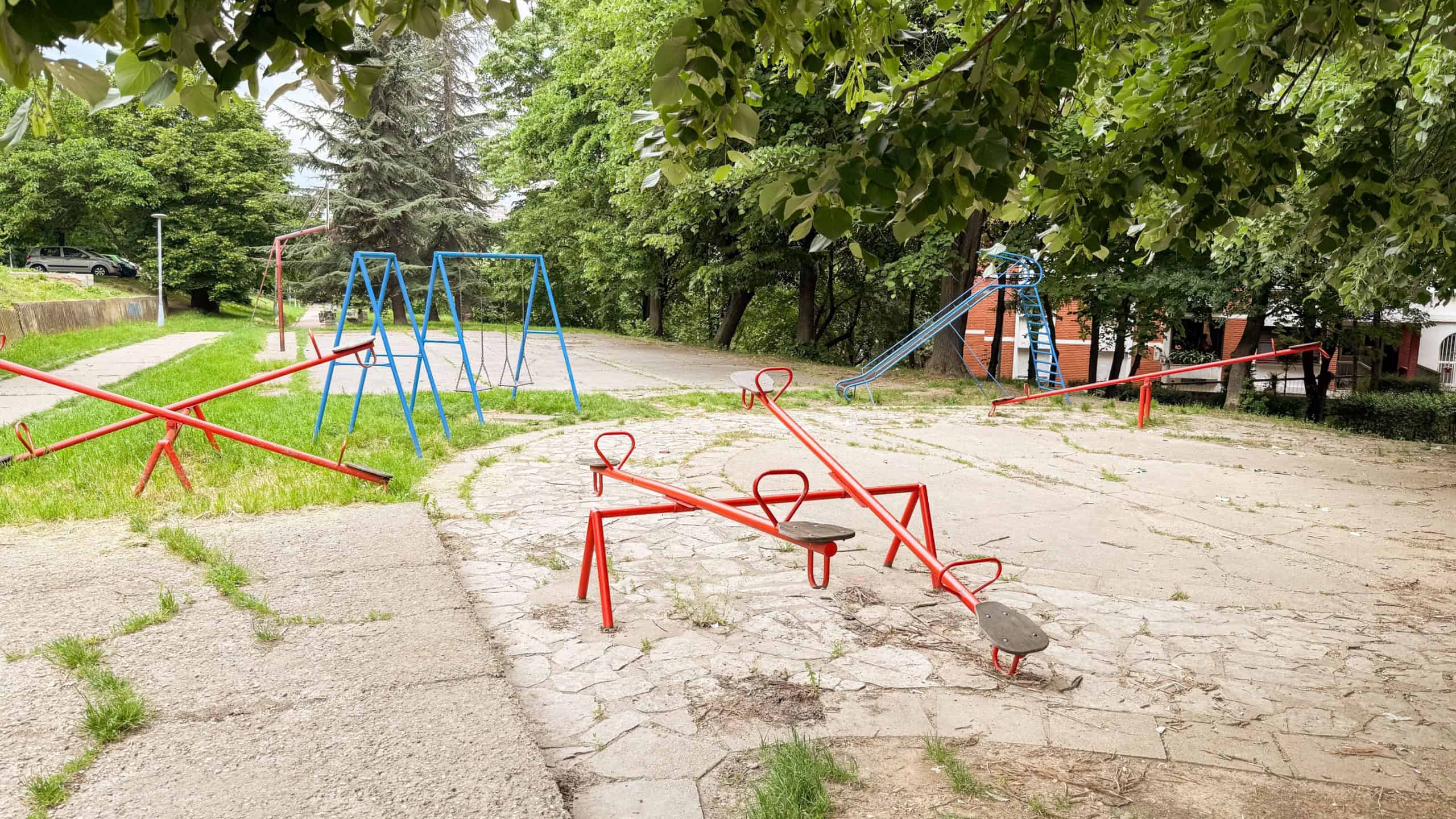 Empty retro playground with seesaws, swings, and a slide under green trees. A quiet and nostalgic public space in the city.