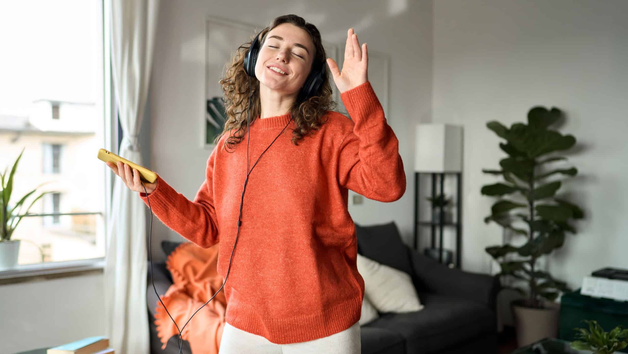 Candid photo of happy pretty young woman wearing headphones dancing at home. Cheerful girl listening music on mobile phone, singing song feeling relaxed standing in modern living room interior.