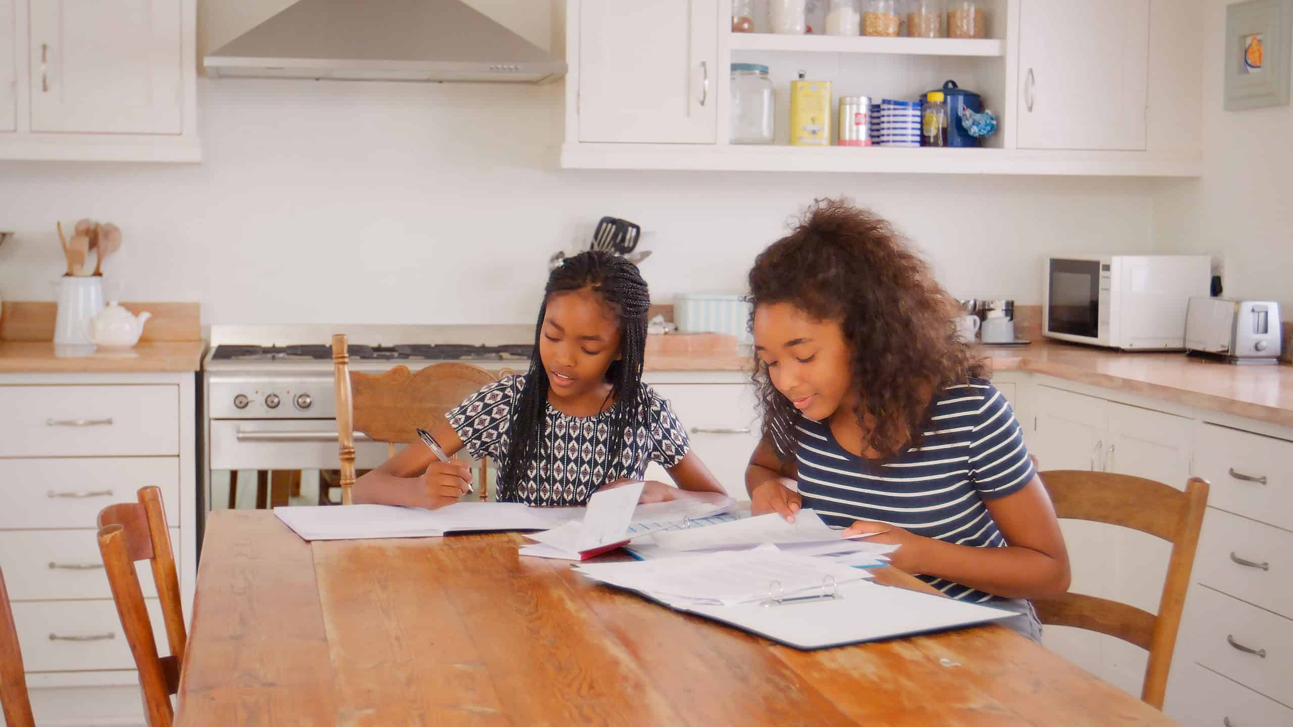 Teenage Girl Helping Sister With Homework Sitting At Table In Kitchen At Home