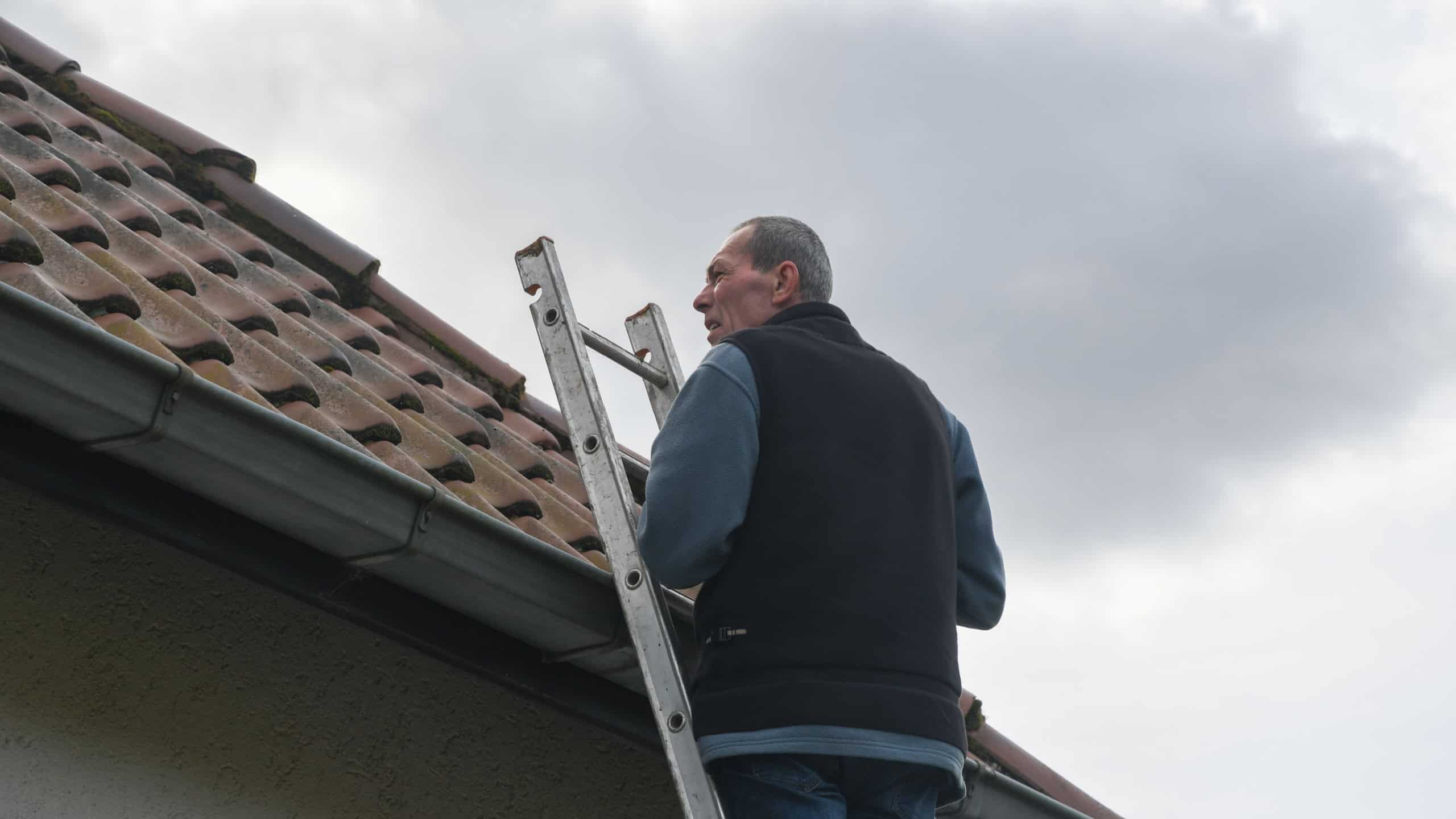 Senior roofer climbs an aluminum ladder, carefully inspecting the roof of a residential building under a cloudy sky, taking precautions during maintenance,