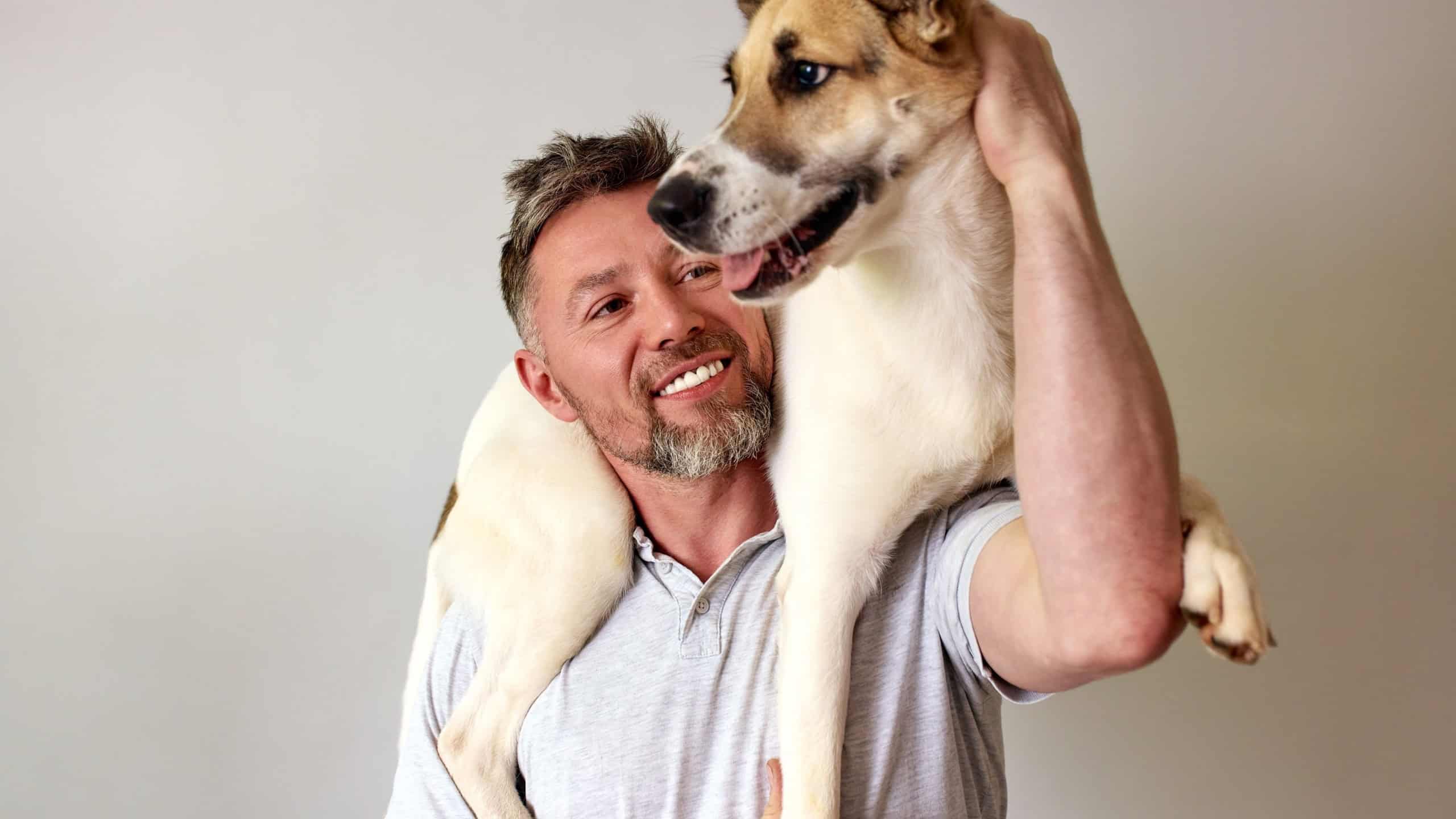 Smiling man joyfully holding his dog over his shoulder in a light room