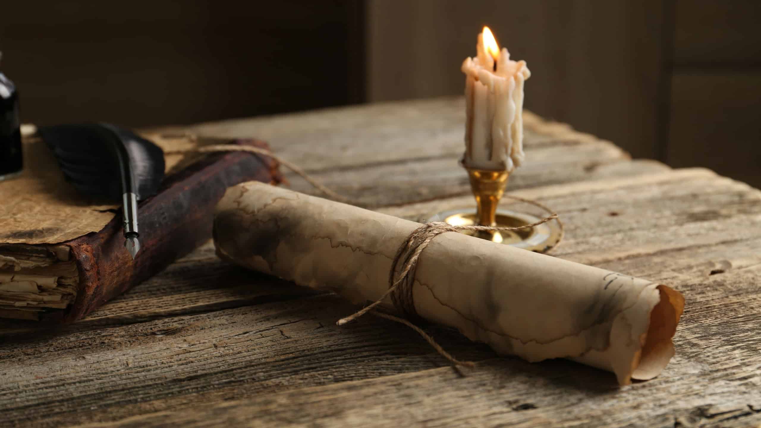 Old paper scroll, book, feather and burning candle on wooden table, closeup