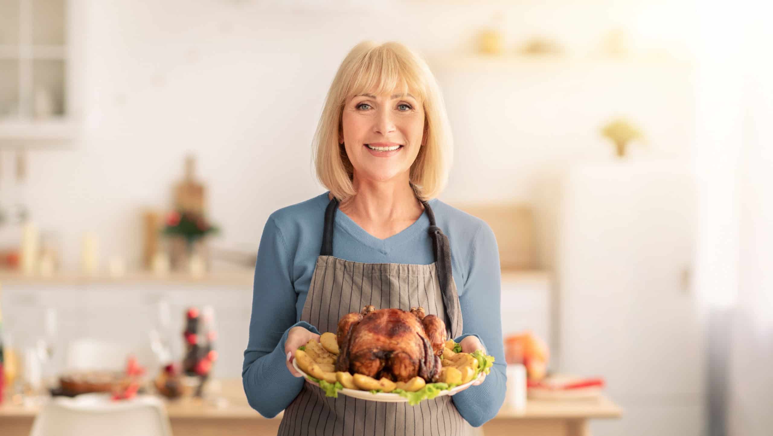 Cheerful elderly woman in apron holding dish with roasted turkey for Christmas or Thanksgiving holiday in kitchen