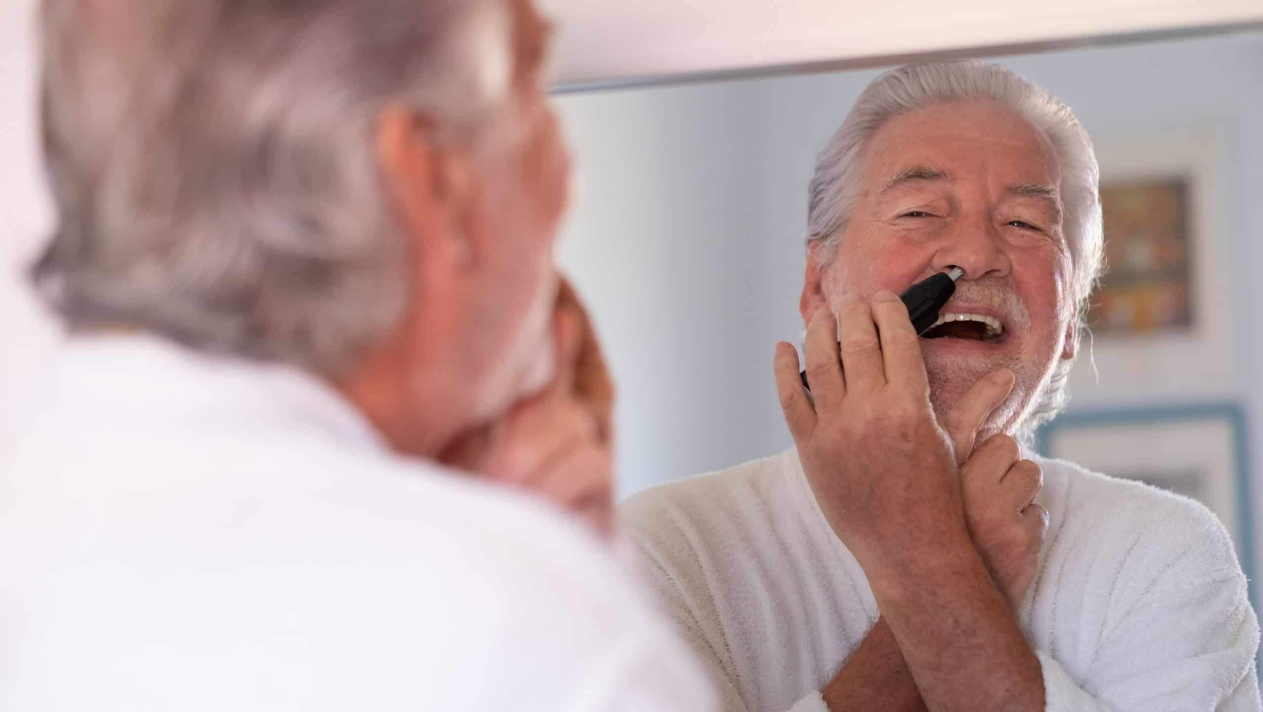 Portrait of senior bearded man in bathrobe while remove nose ear hair with help of little comfy trimmer looking at himself in the mirror