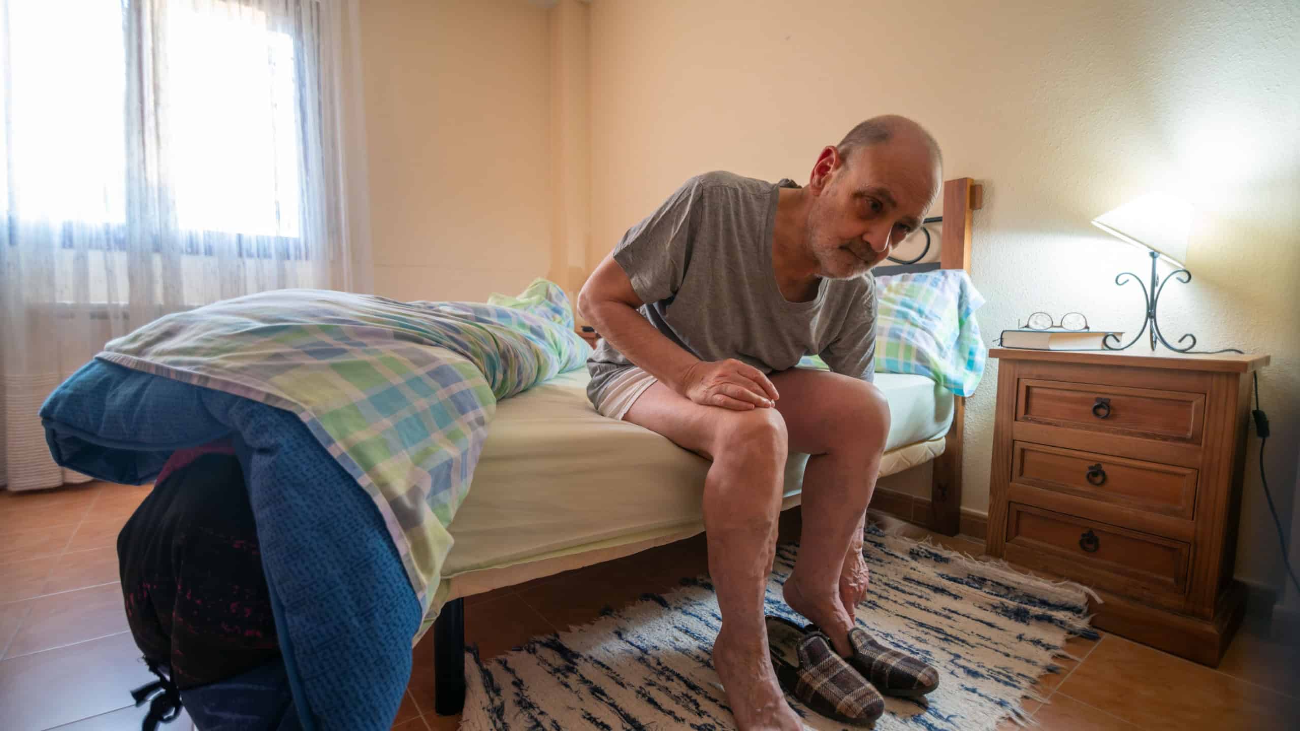A senior man sits on the edge of a neatly made bed in a cozy bedroom. Barefoot with slippers on the floor, he looks thoughtful as morning light fills the room beside a nightstand and lamp