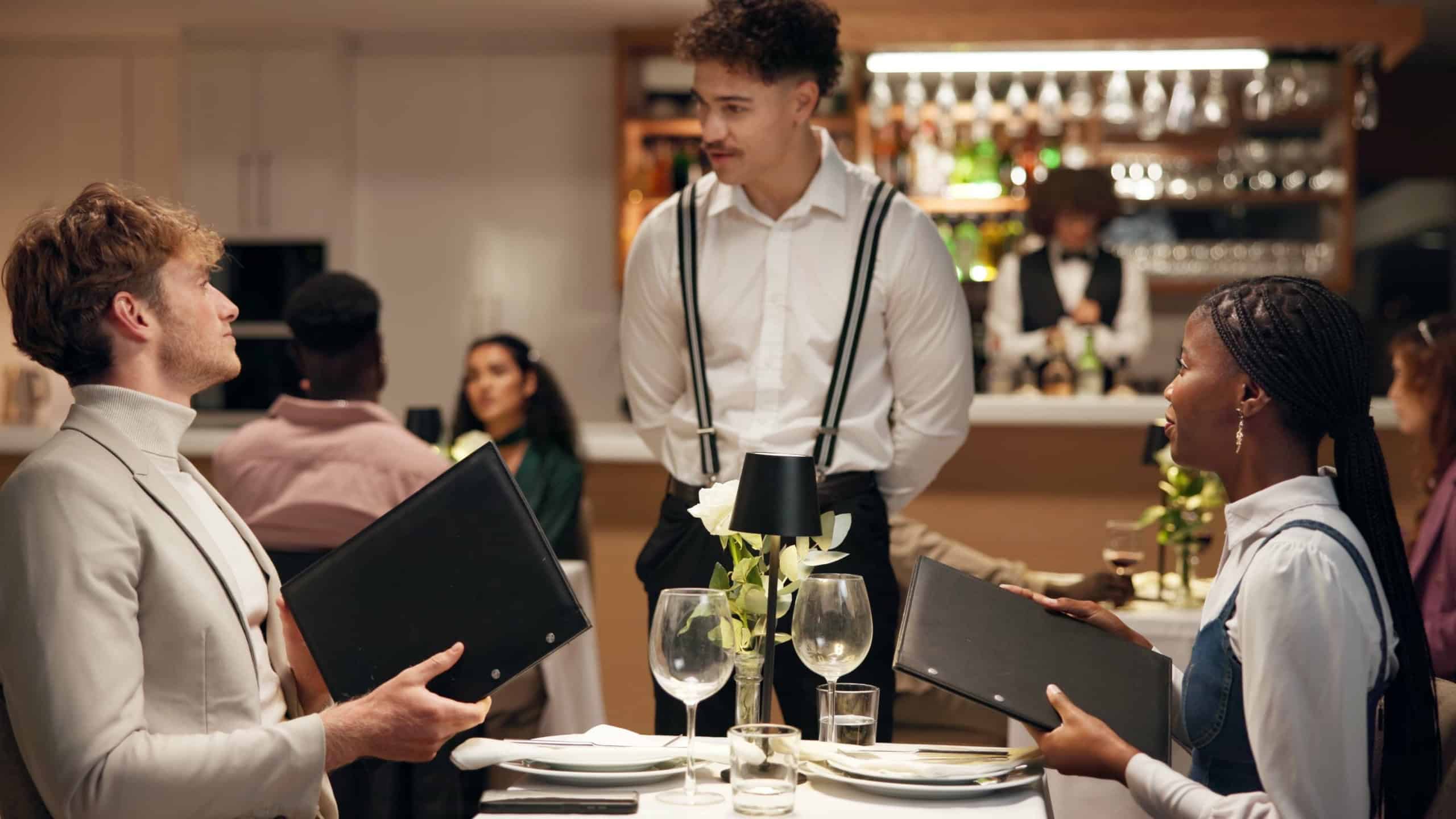 Waiter, couple and talk at restaurant with menu, decision and discussion for wine list, food or order. People, server and customer experience with service, question and opinion for choice at dinner