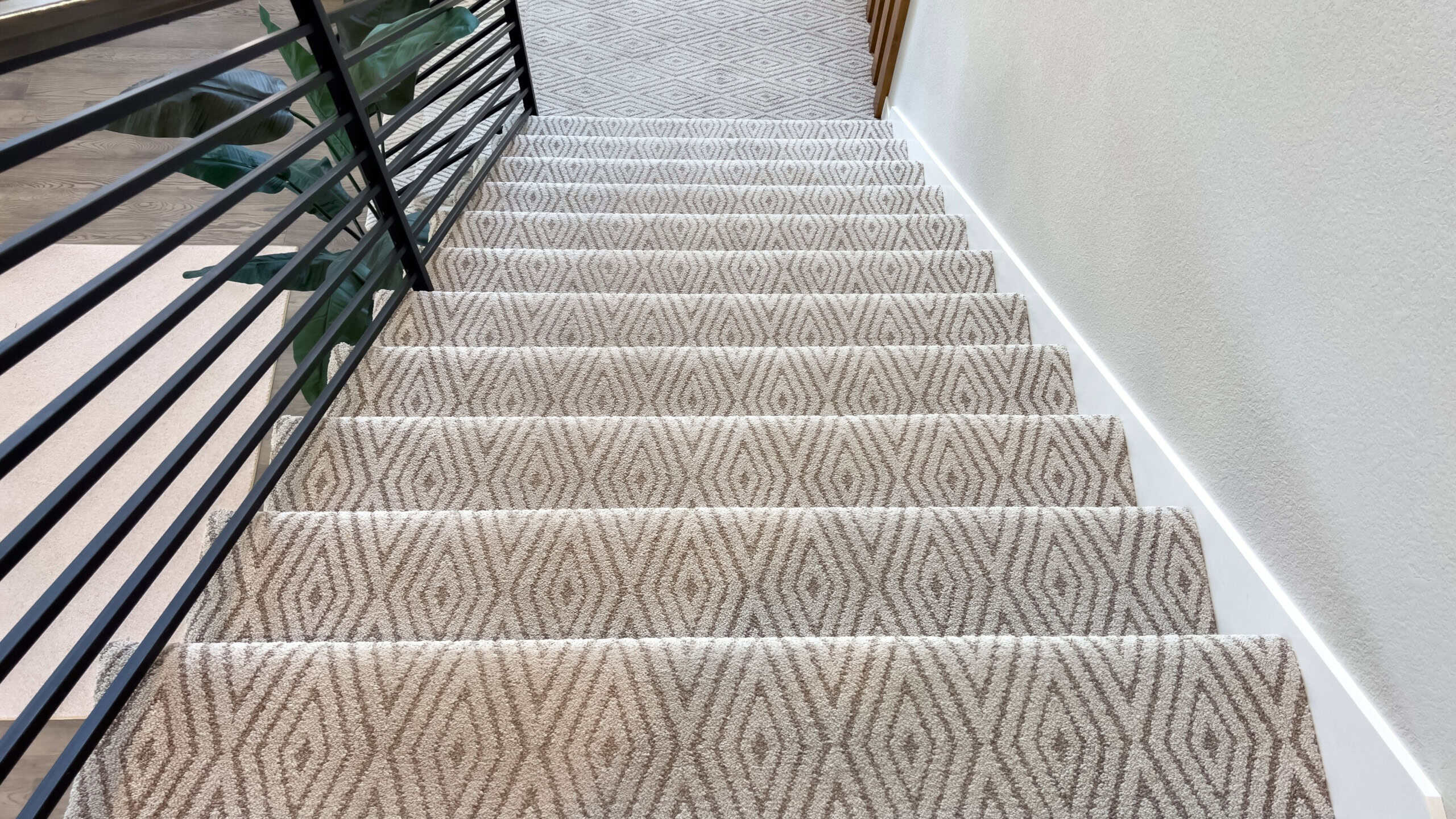 A top-down view of stairs with a geometric carpet runner and black metal railing. The modern design highlights symmetry, pattern, and structural contrast.