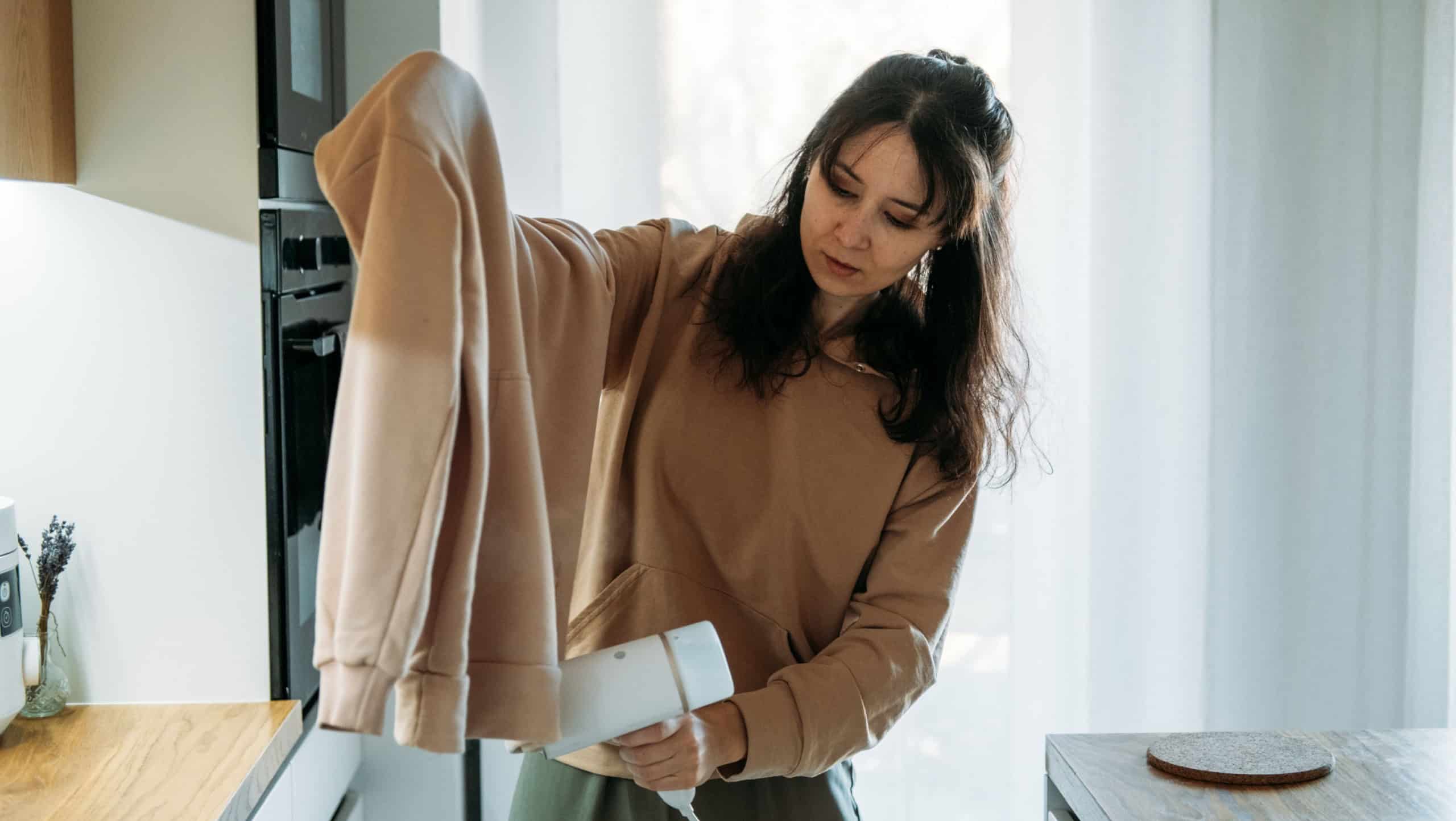 Woman uses portable handheld steamer on sweatshirt in modern kitchen. Time saving technology, modern laundry care, domestic efficiency.