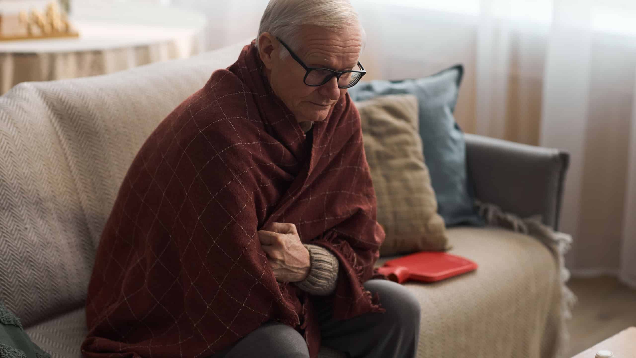 Senior Caucasian man sitting on sofa wrapped in blanket, holding arms close to chest, appearing cold or unwell, wearing glasses, looking down with thoughtful expression of health concern