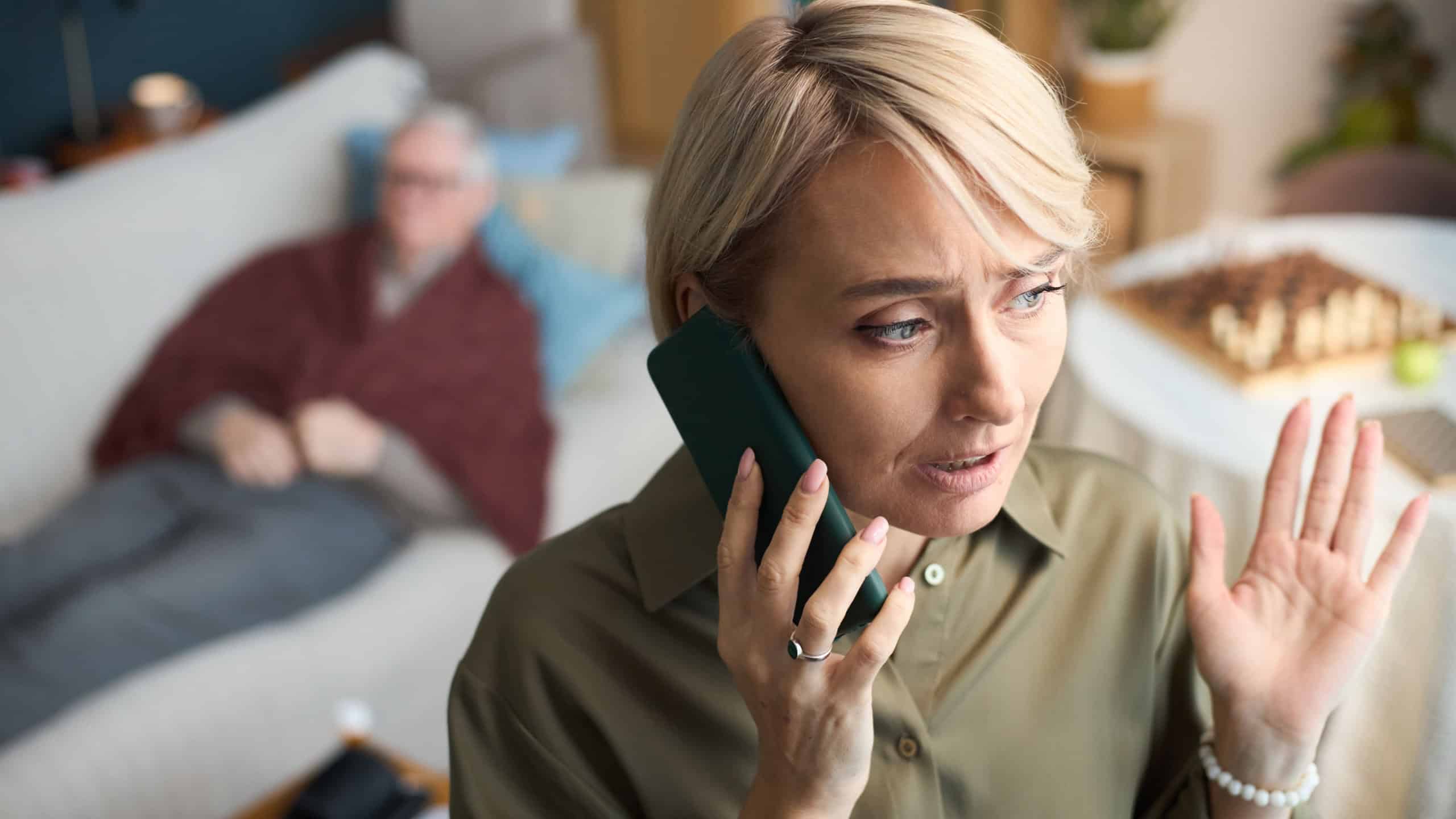Caucasian middle aged woman talking on smartphone with concerned expression, calling for doctor for sick father, raising hand while sitting in living room, senior man resting on sofa in background