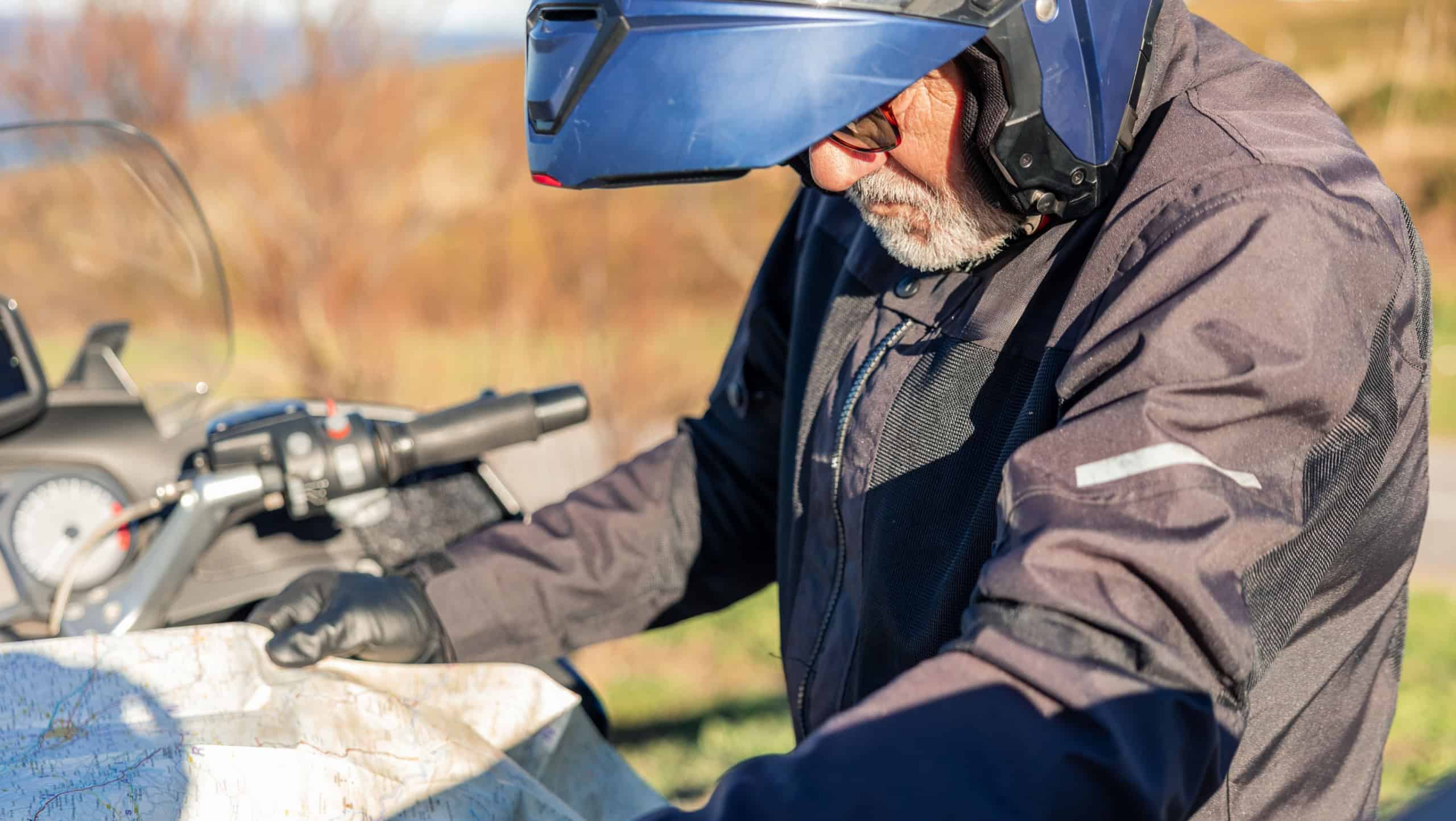 Elderly man wearing a helmet and jacket looking closely at a paper map, planning his journey while stopped on his motorcycle on a sunny day during an outdoor adventure