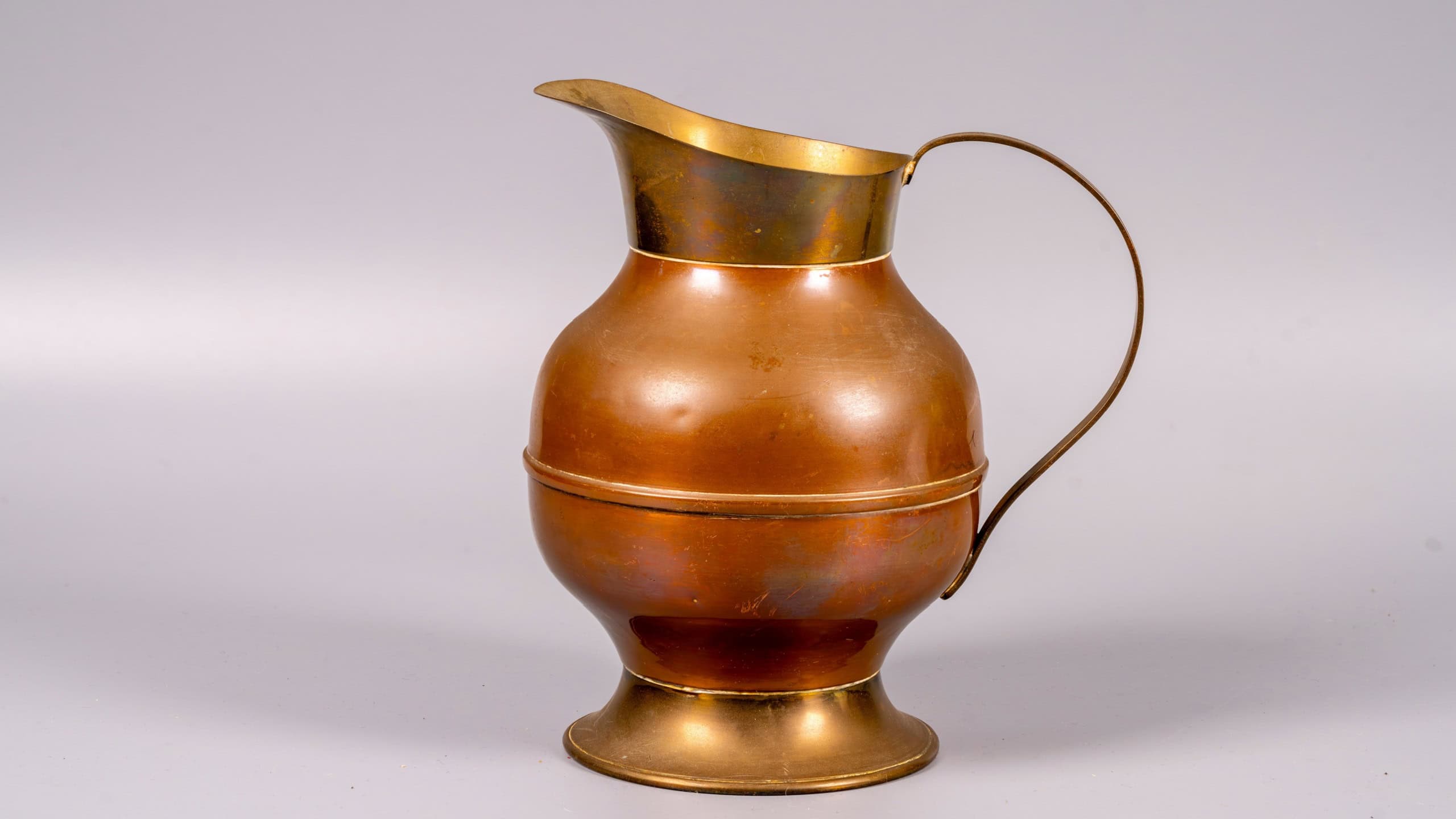 A copper pitcher is placed on a plain background. The pitcher has a rounded body and a long spout. It stands on a wide base and is designed for pouring liquids.
