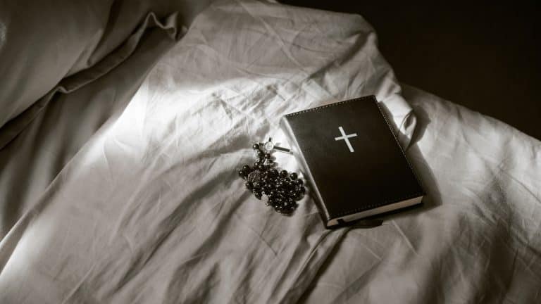 Closed Bible with cross symbol and rosary beads resting on bedsheet, religious objects positioned together on bed, symbolizing Christian faith and devotion
