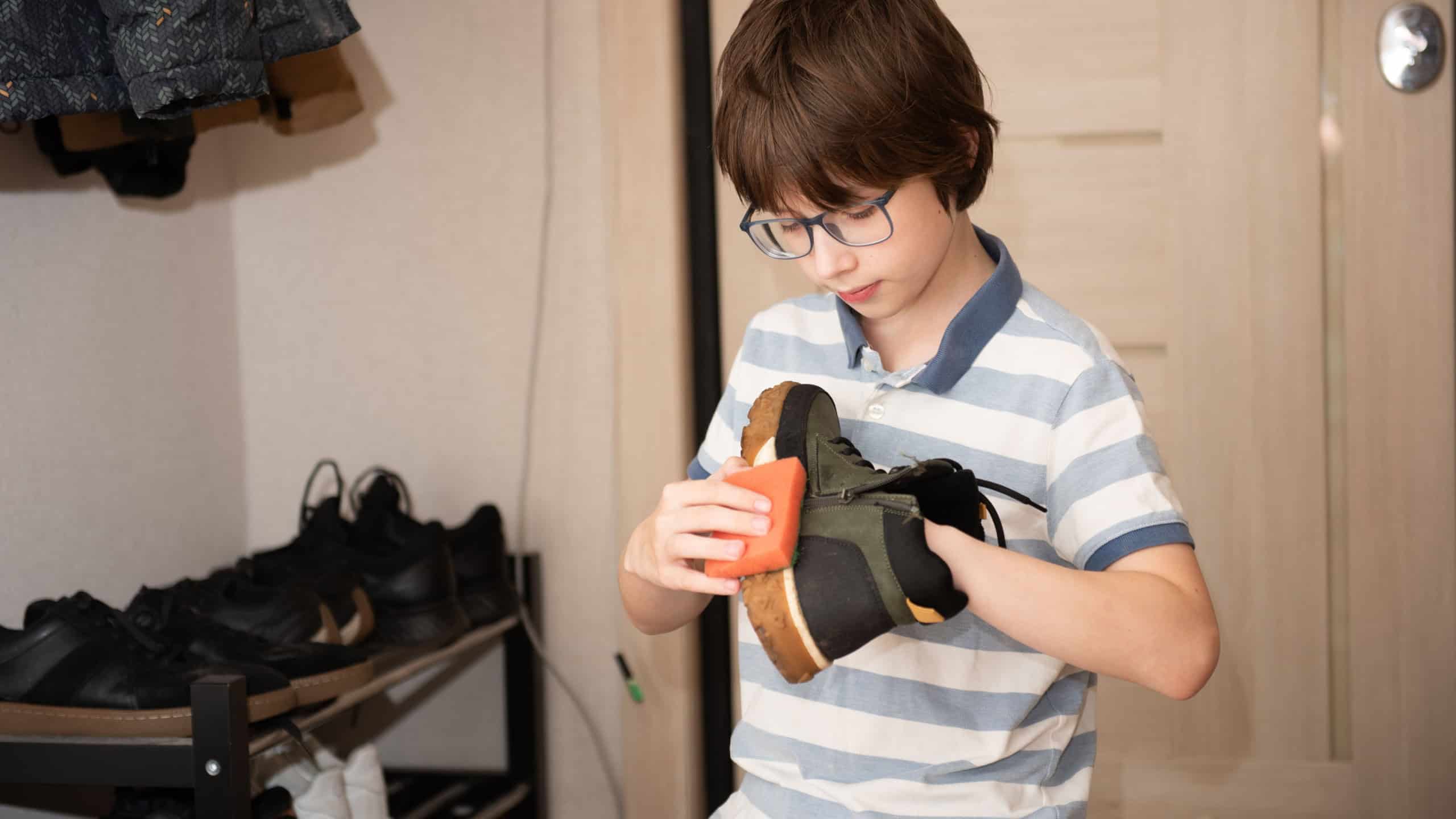 Young boy cleaning a shoe with a sponge in a home hallway.