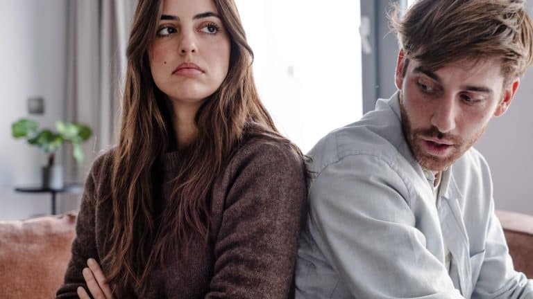 Young couple sitting back to back on couch having a dispute, expressing relationship conflict, unresolved problem or question, emotional distance, or communication problems in a modern living room.