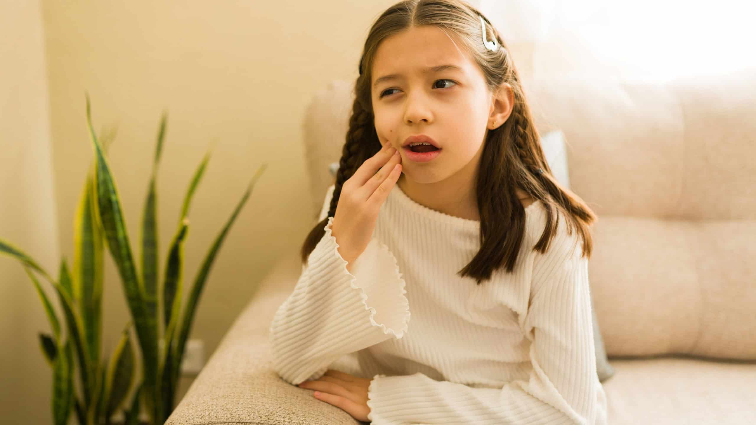 Young girl experiencing strong tooth pain, touching her cheek, sitting on sofa, needing immediate dental attention