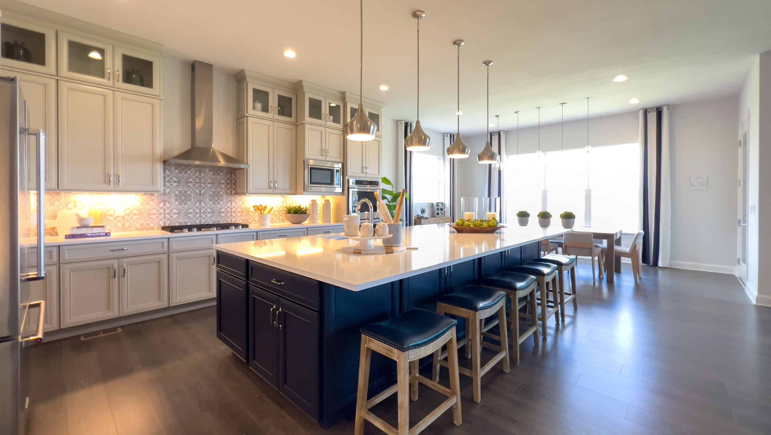 A full view of a contemporary kitchen with dark lower cabinets and light upper cabinets, a wide island with bar stools, and pendant lighting. Dining space visible in the background.