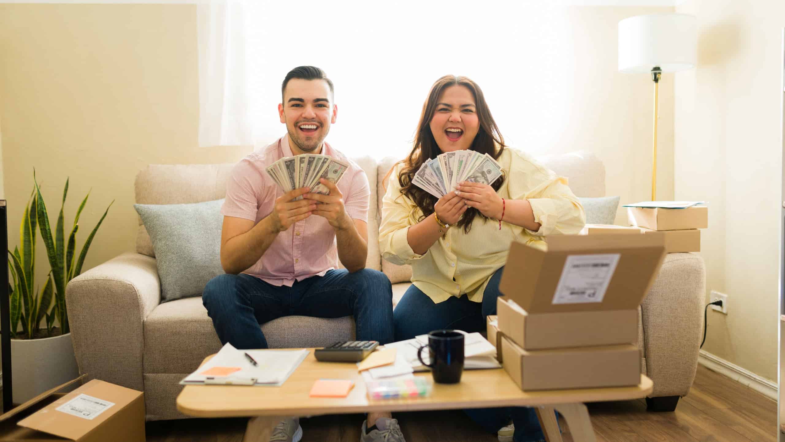 Cheerful couple holding dollar bills, celebrating their profitable side hustle, packaging orders for shipment, achieving financial success