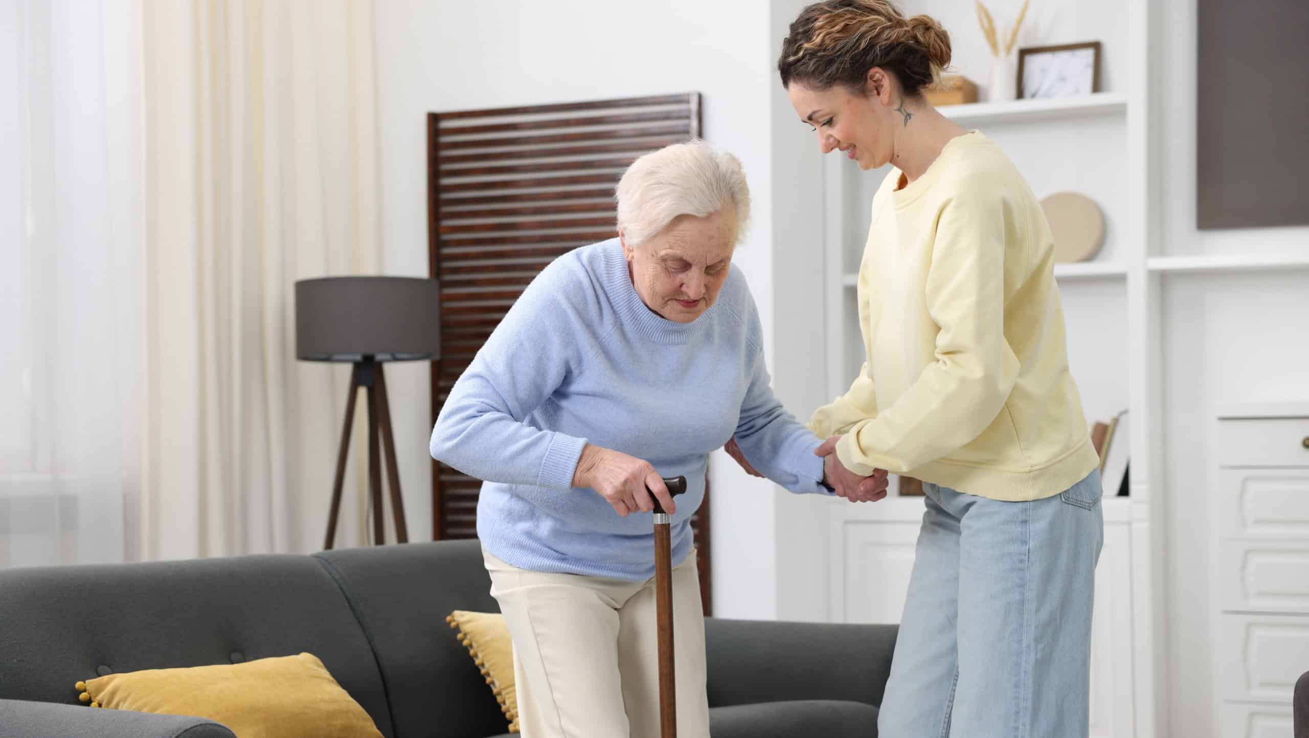Care worker helping senior woman with walking cane to stand up from sofa indoors