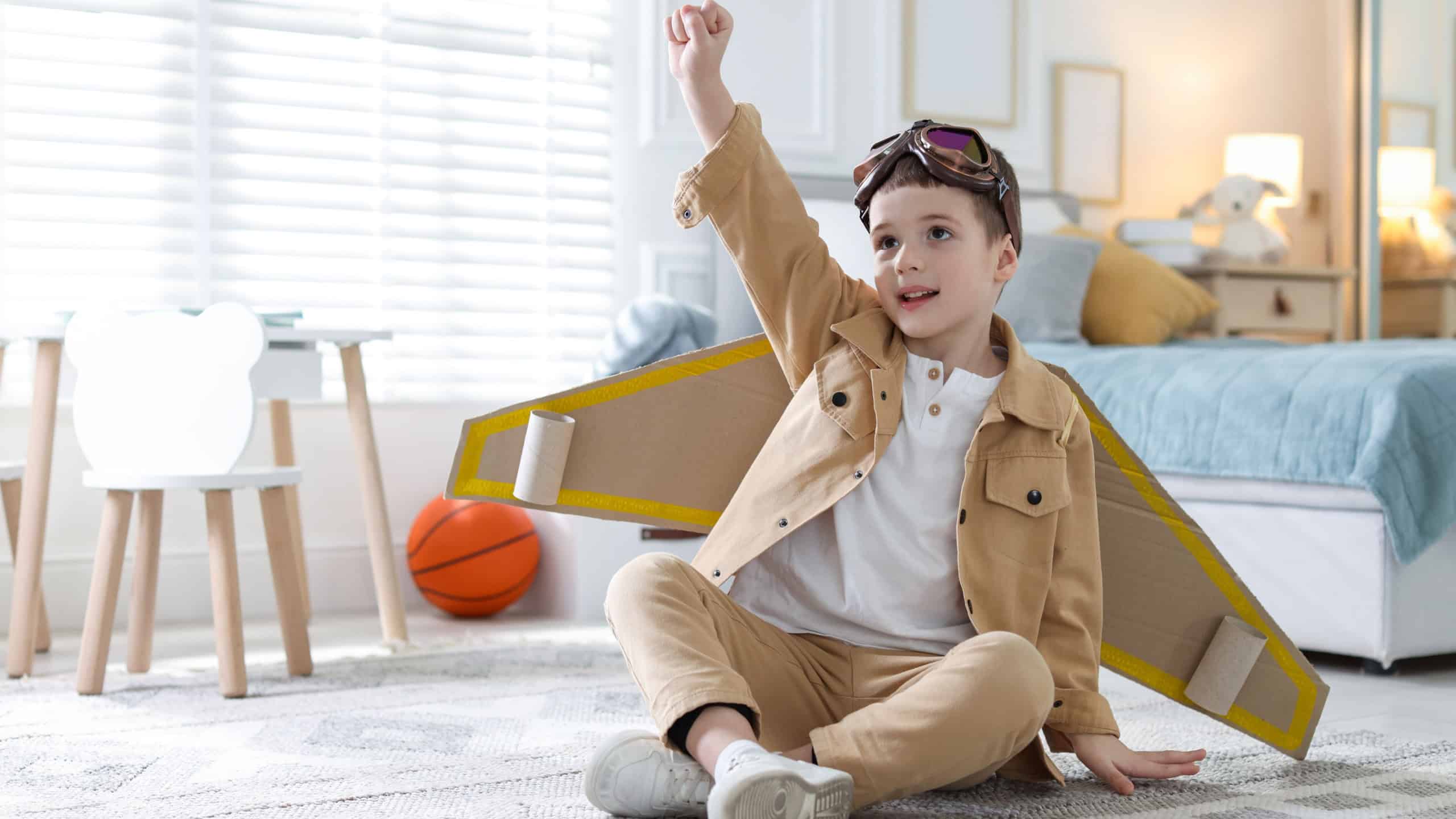 Smiling little boy with cardboard plane wings and goggles playing pilot on floor at home.