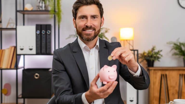 Middle-aged businessman at home office drops coins into piggy bank and plans budget for future goals. Freelancer guy at table saves money counts change and sets investment target for personal finance.