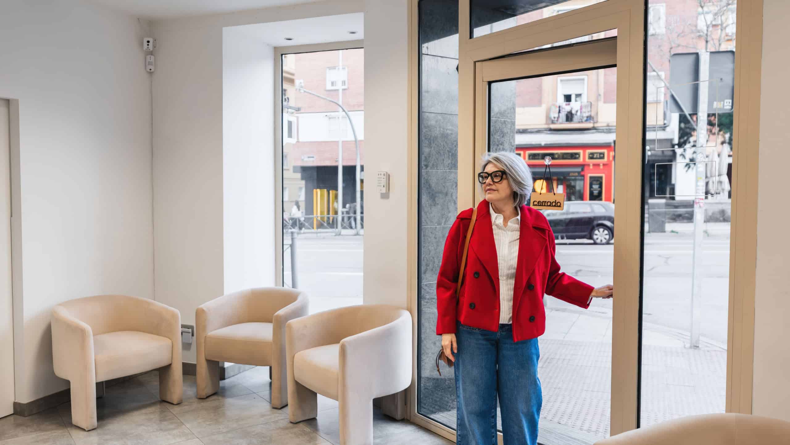 Senior woman in red jacket opening a glass door, entering a modern dental clinic or medical office waiting room