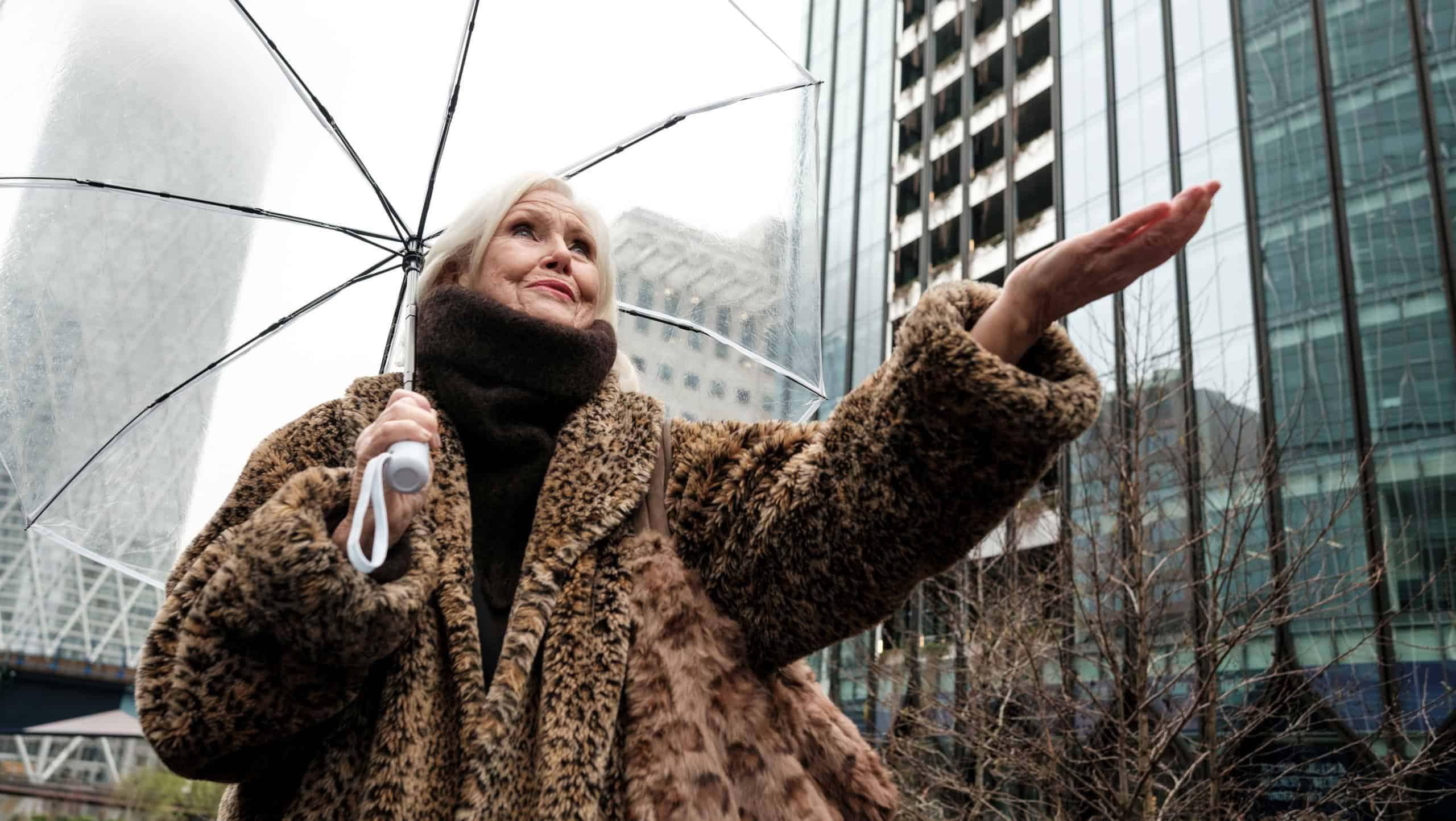 Senior woman wearing a leopard print faux fur coat and warm scarf, holding a clear umbrella while reaching out to feel the falling rain in the city with modern skyscrapers