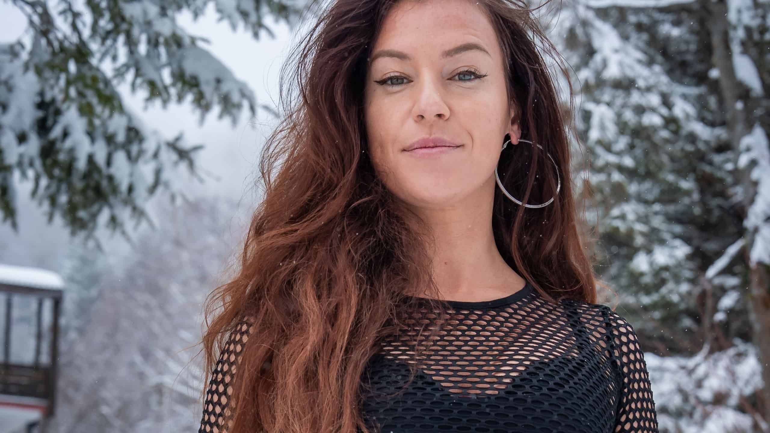 A woman with auburn hair stands on a snowy path at San Domenico, Italian Alps. She wears a black mesh top and hoops as fine snow falls near a chalet in soft light.