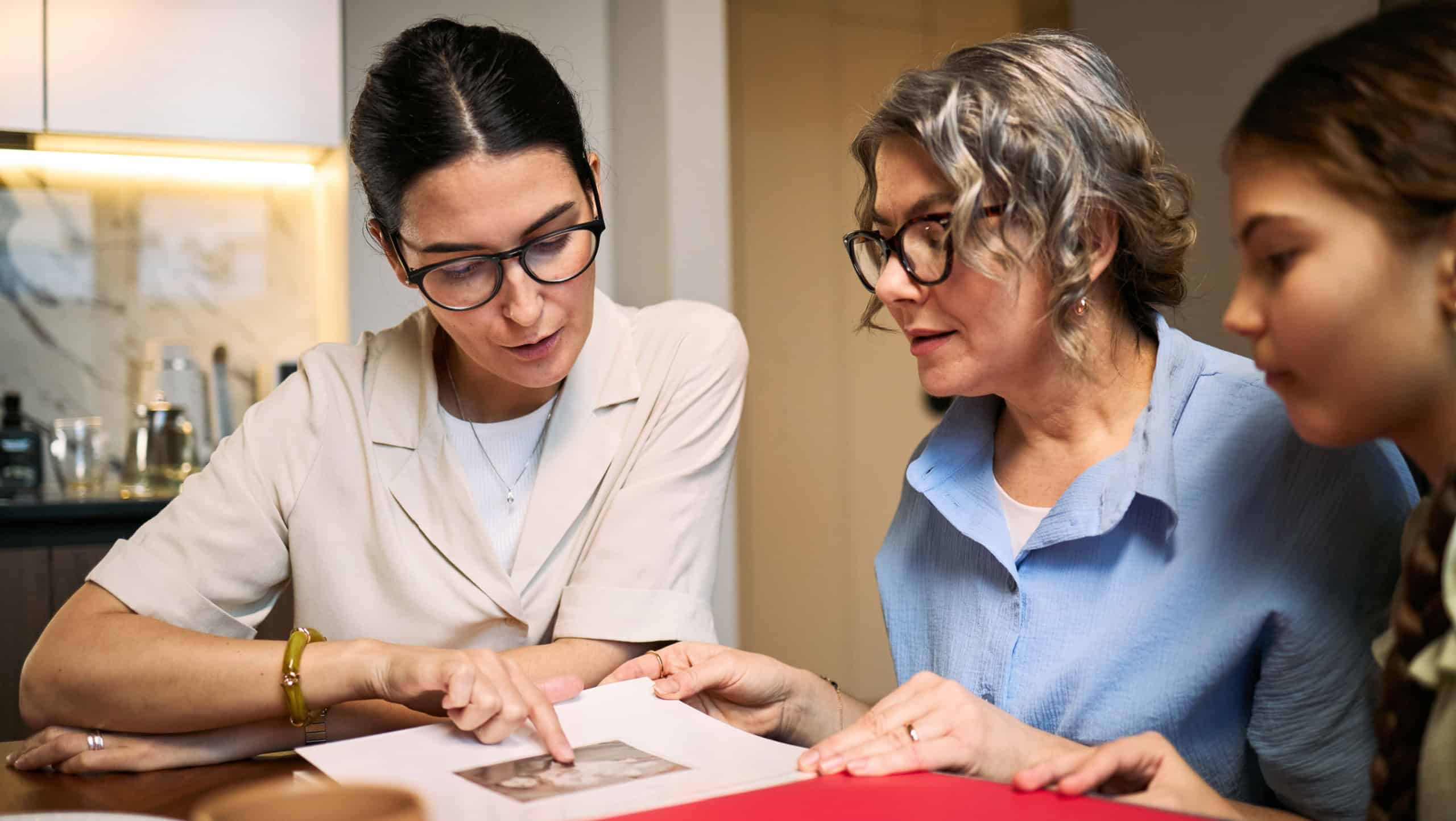 Young adult woman guiding middle aged woman and girl while reviewing family photo album at table. Suitable for family history, caregiving support, memory preservation marketing, medium shot