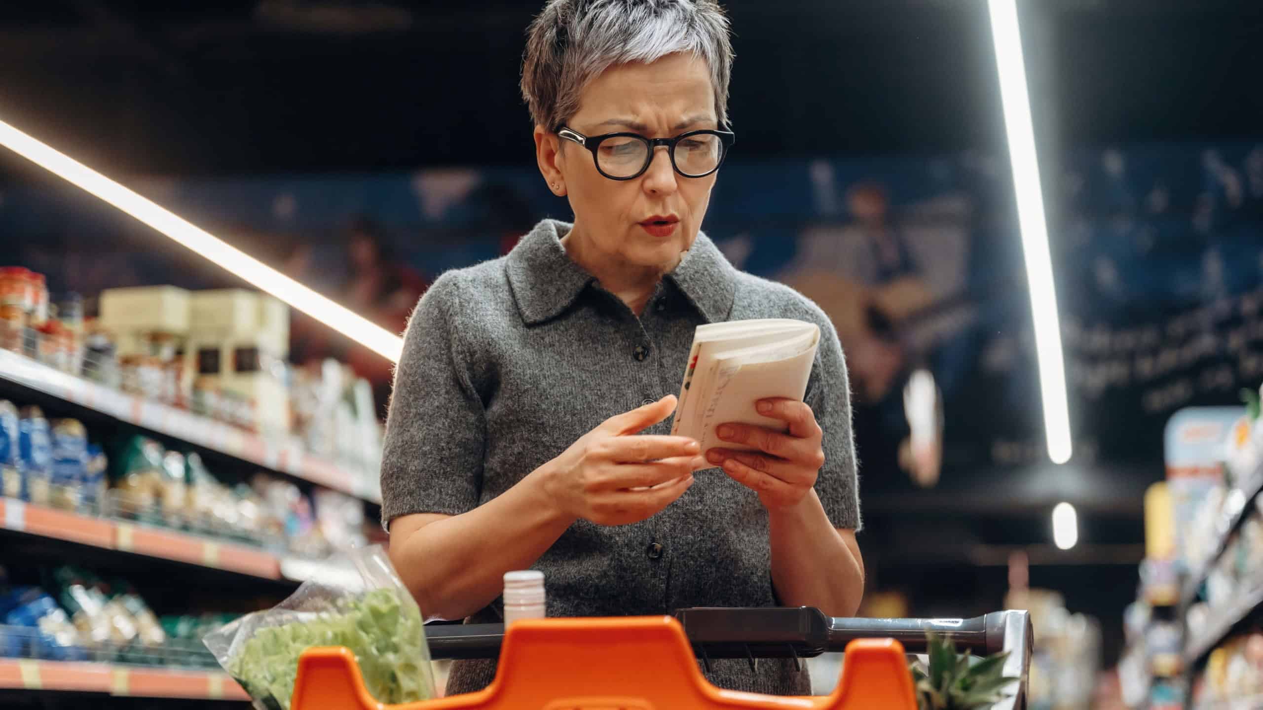 Mature Asian woman reads a product label while standing in a grocery store aisle, surrounded by shelves stocked with food items and colorful packaging