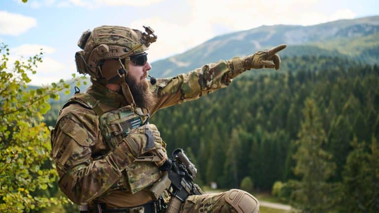 Bearded Soldier in Tactical Gear Pointing Toward Distant Mountains During Forest Reconnaissance Patrol