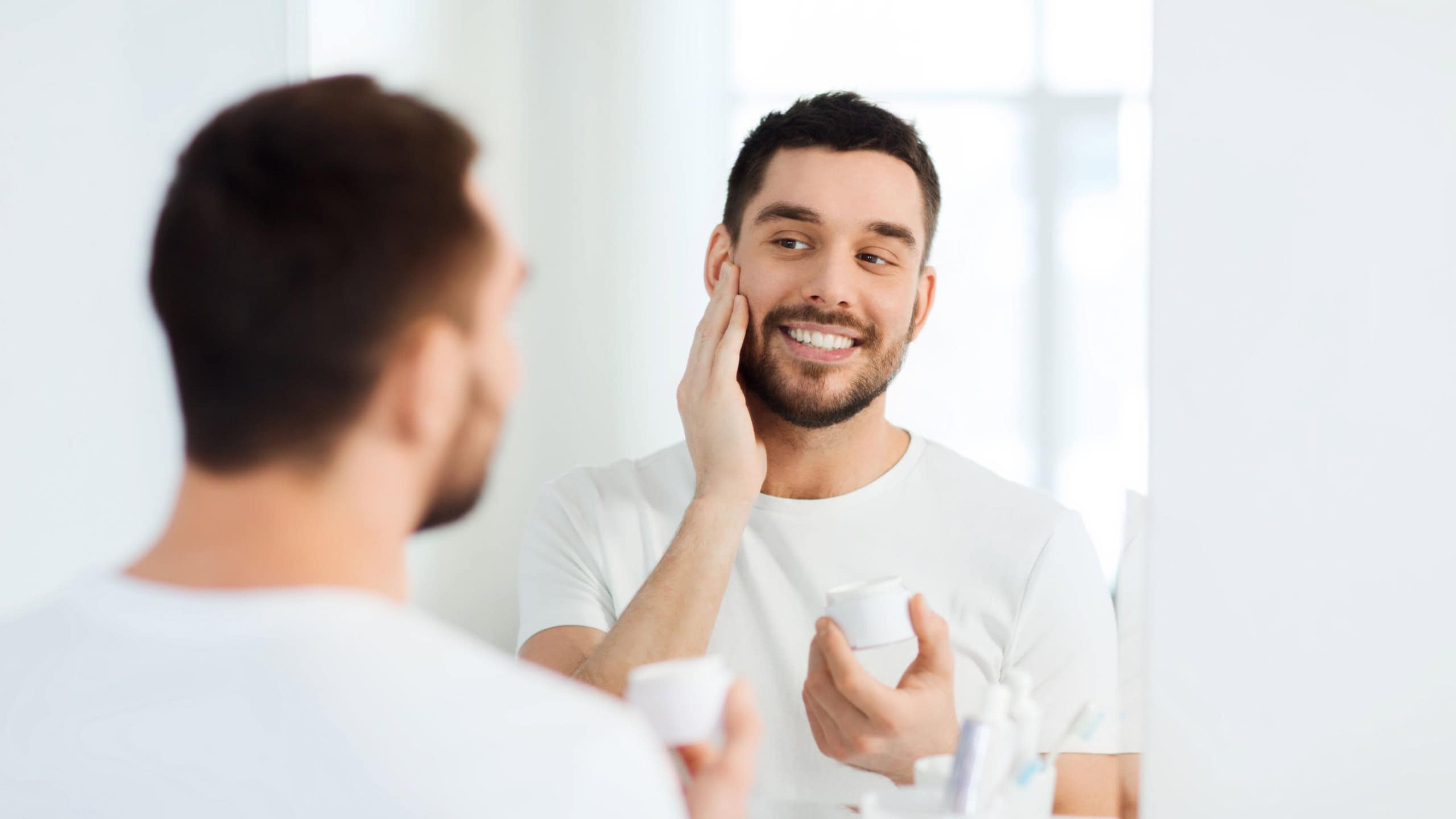 Beauty, skin care and people concept - smiling young man applying cream to face and looking to mirror at home bathroom