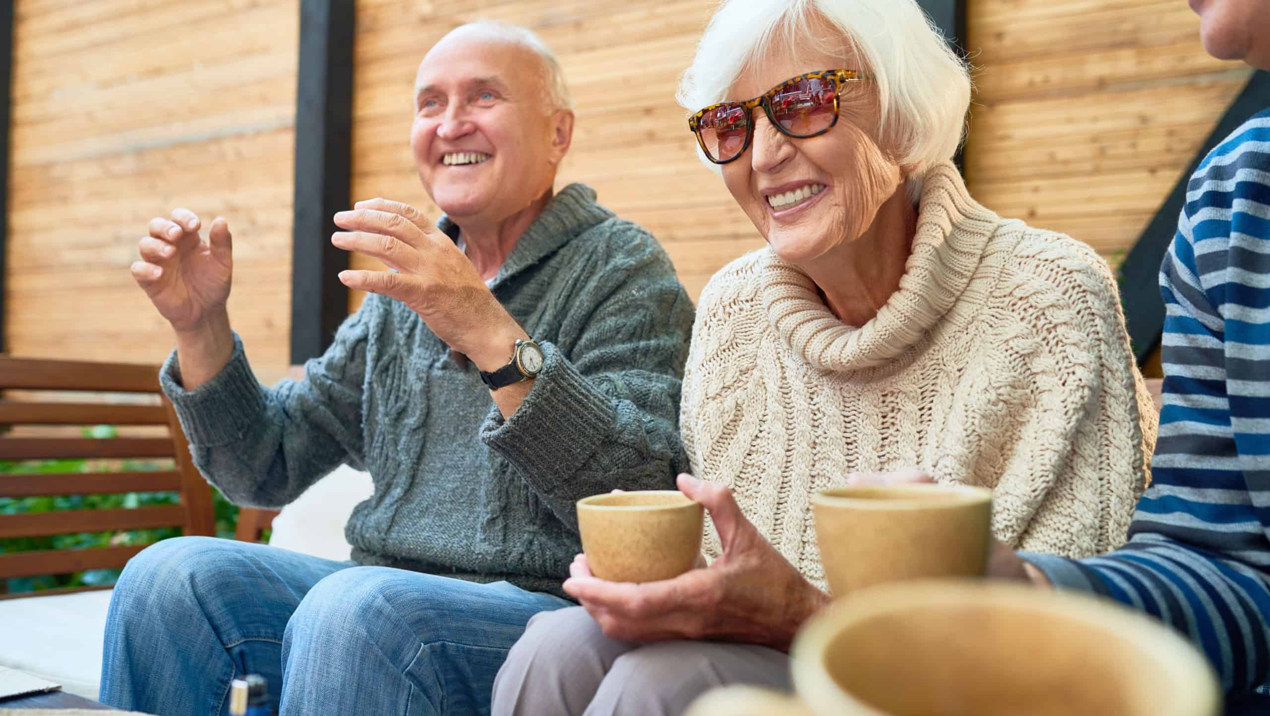 Cheerful senior friends gathered together at cozy small patio and remembering funny stories from their past, they wearing knitted sweaters and warming themselves with herbal tea
