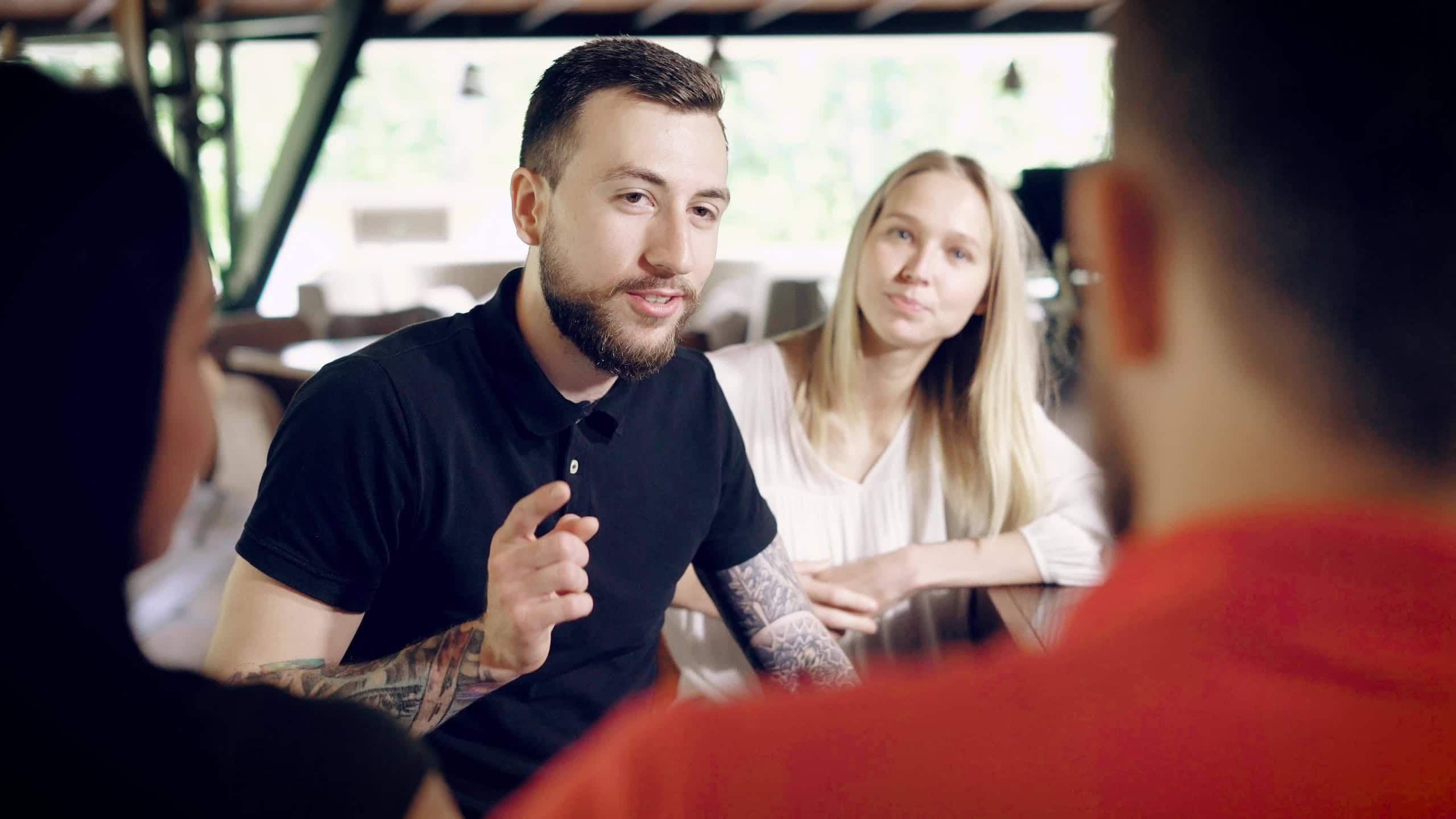 A stylish and bearded man with a tattoo on his hand communicates with friends in a bar or restaurant, a man tells a funny story