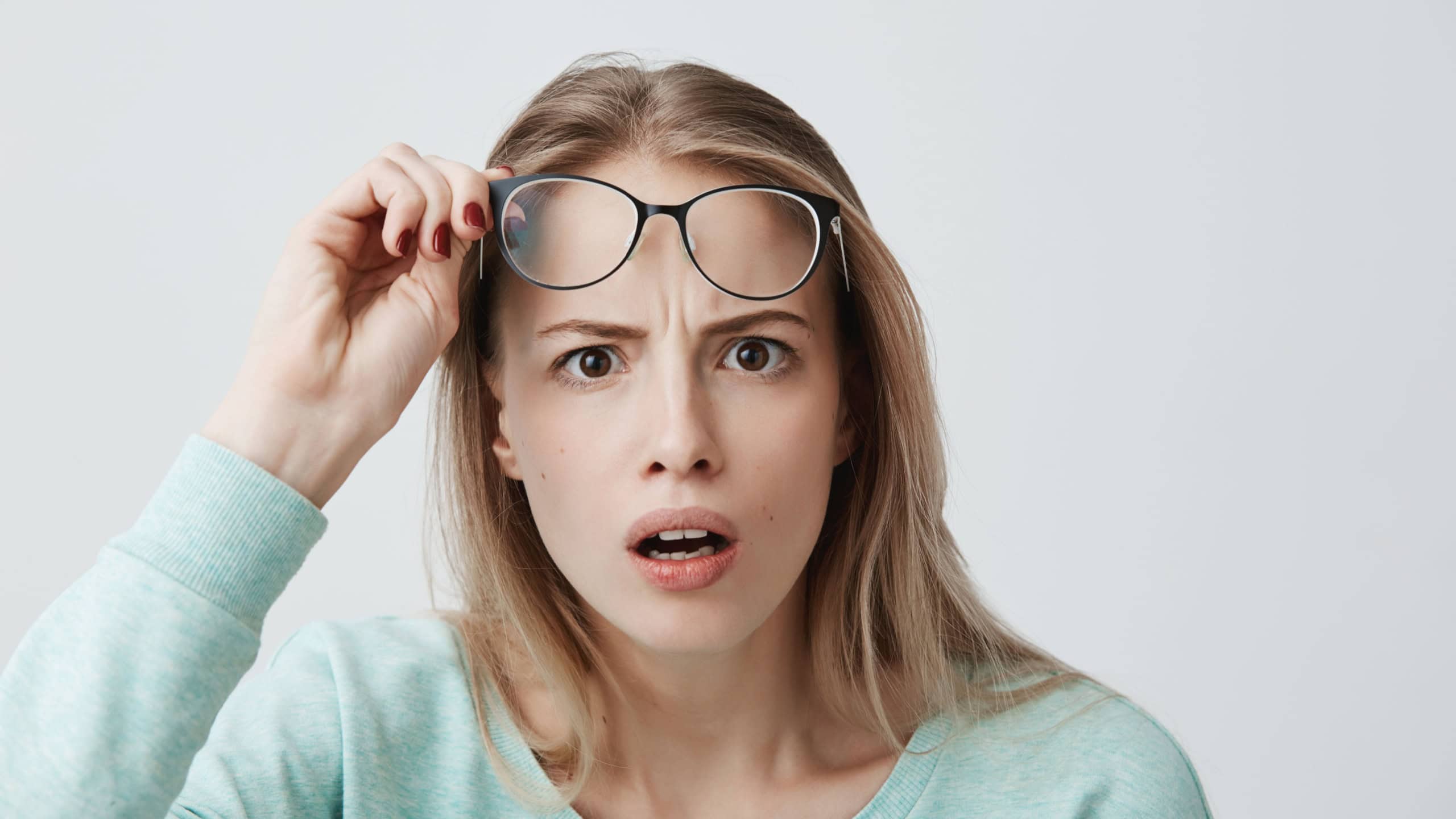 Surprised young female model with long blonde hair, wears glasses and blue long-sleeved shirt, looks with terror at camera as notices something unexpected, hears bad news, isolated against gray wall