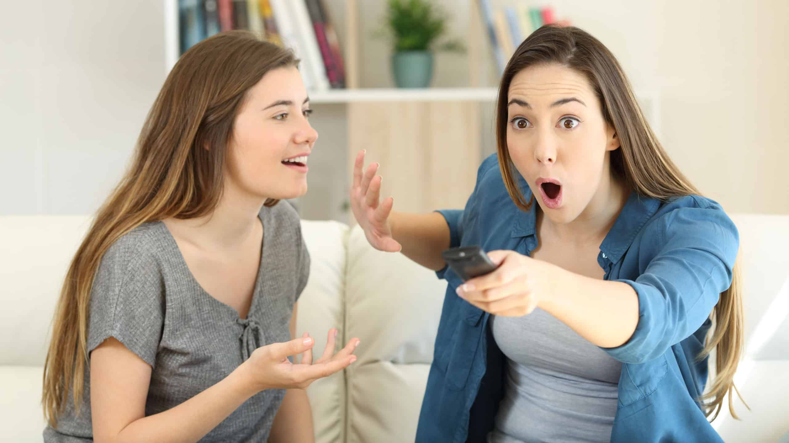 Amazed girl interrupting her friend who is talking to watch tv sitting on a couch in the living room at home