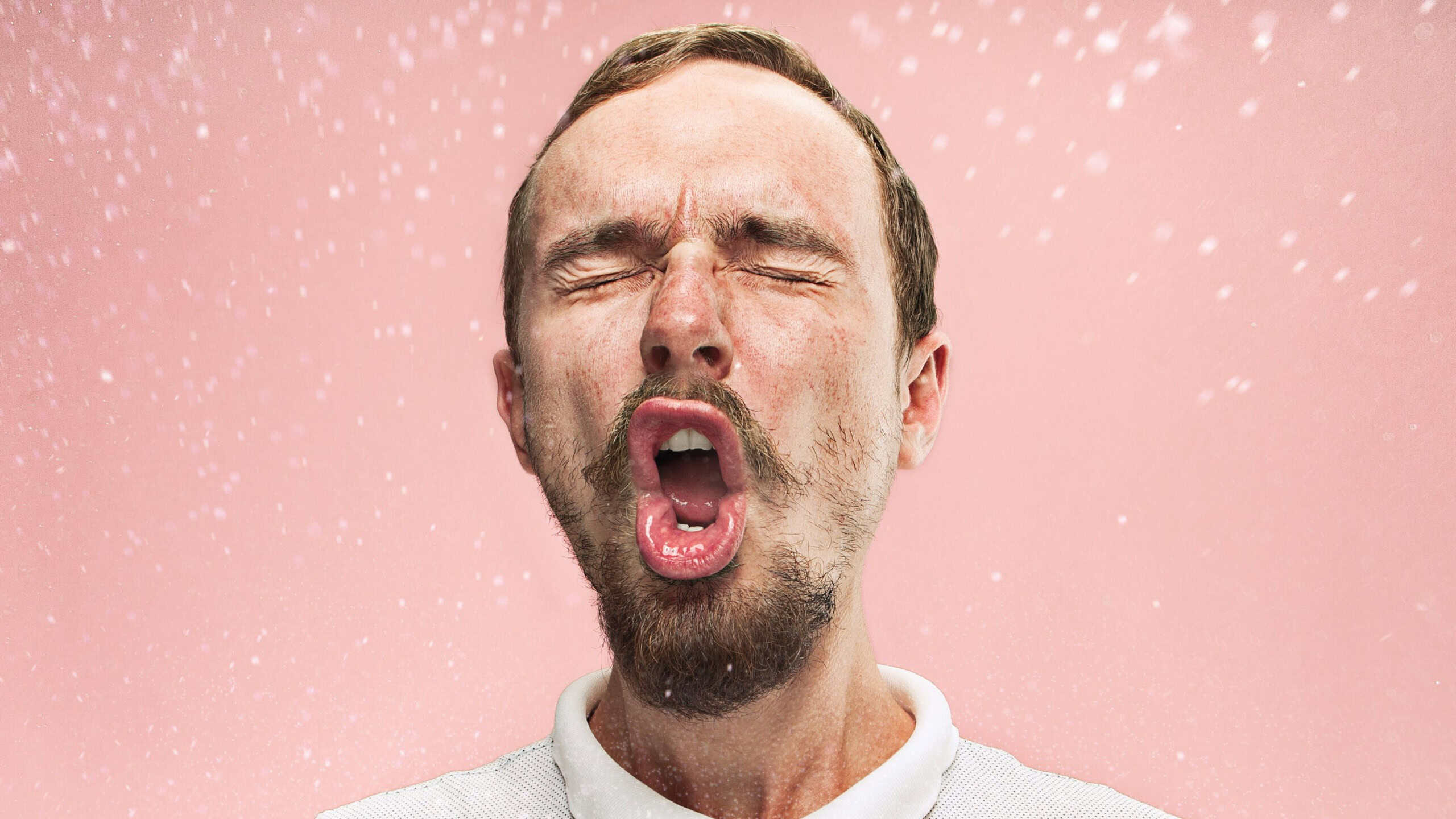 Young funny handsome man with beard and mustache sneezing with spray and small drops, studio portrait on pink background. Comic, caricature, humor. illness, infection, ache. Health concept