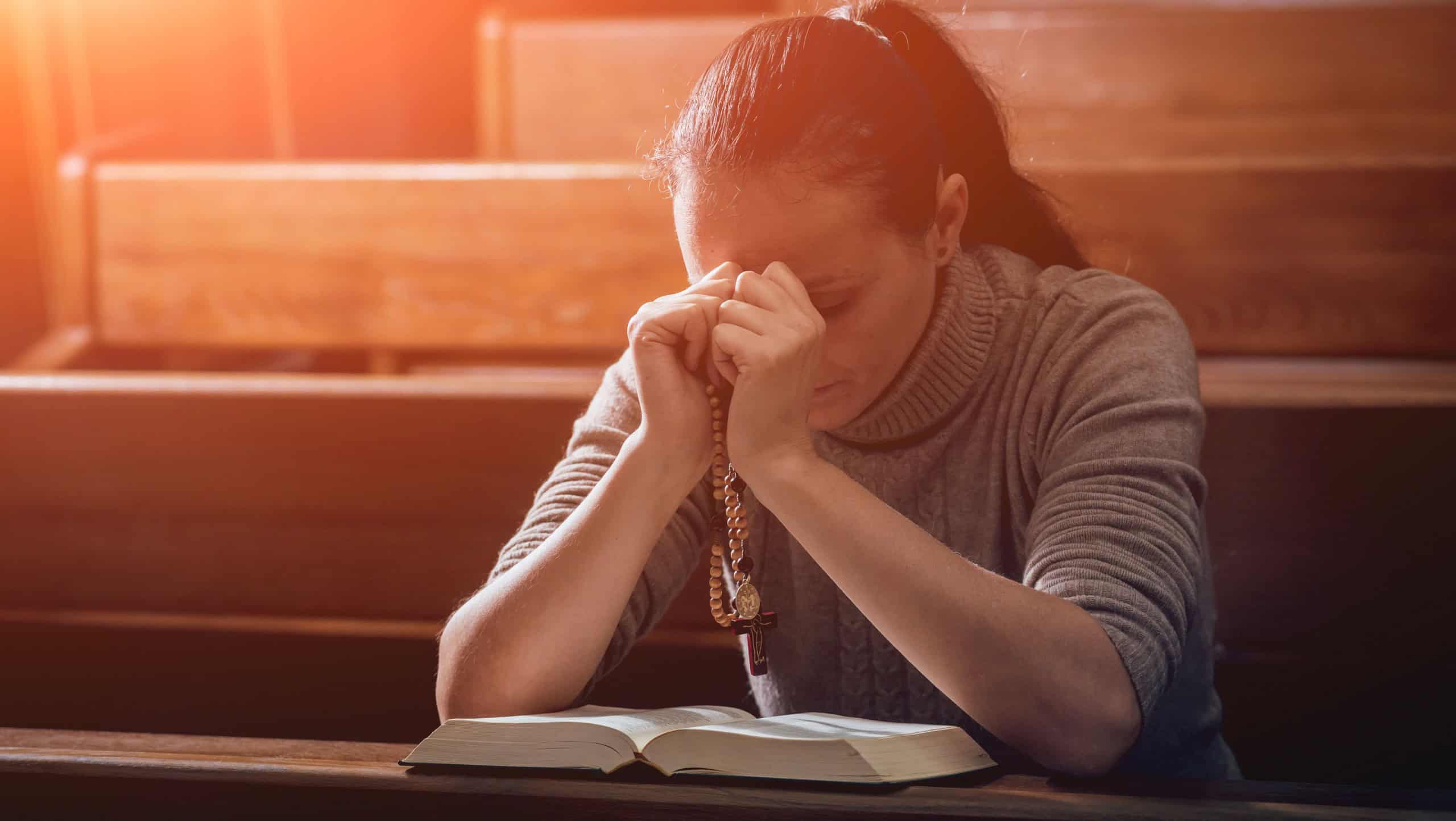 Christian woman praying in church. Hands crossed and Holy Bible on wooden desk. Background