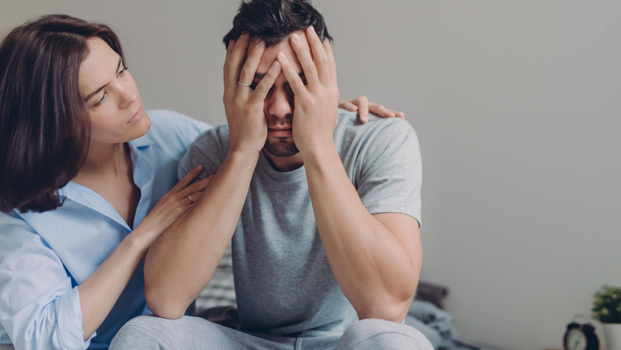 Stressful young man cries from despair, loving mother tries to calm him, pose together at bed against domestic interior, have some problems. Upset husband sees no way out in difficult situation