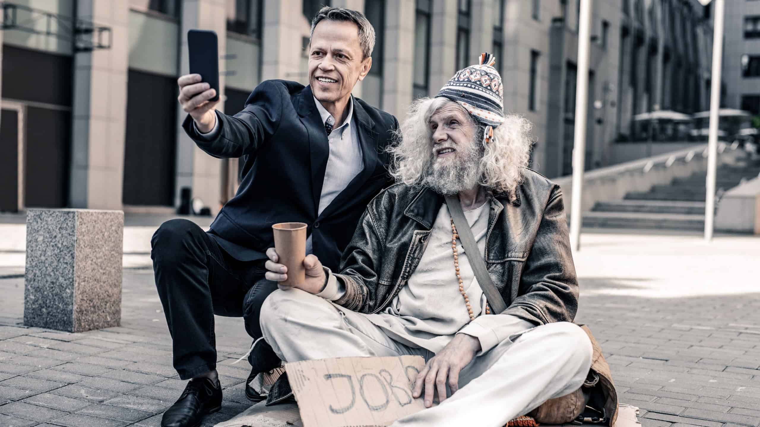 Man taking selfie. Beaming grey-haired homeless looking on the money with wide smile while supporter takes selfie with him